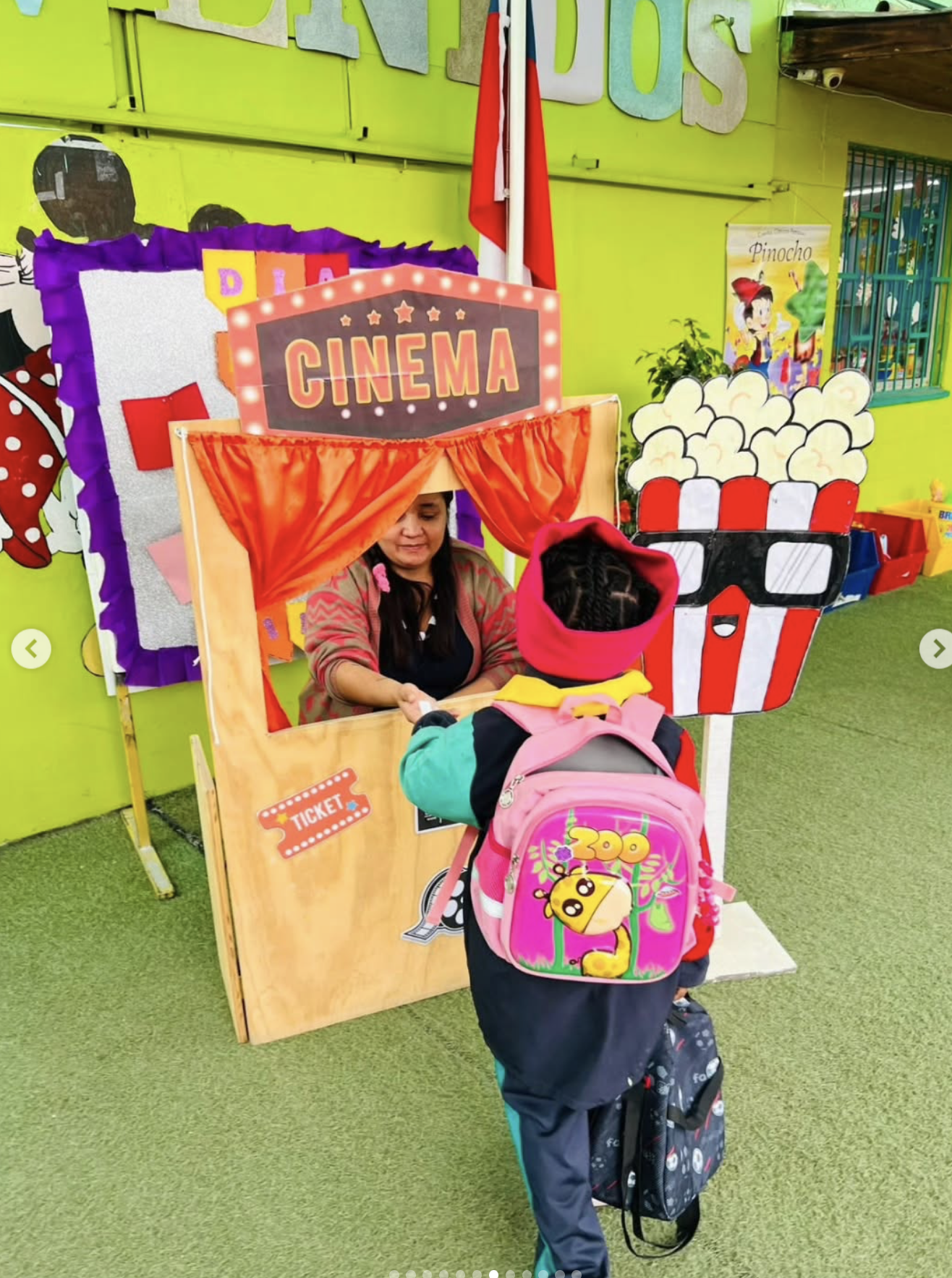 Niña con mochila de color rosa y niño con mochila de color oscuro esperando en una taquilla para comprar boletos de cine en un ambiente colorido con cartel de película de Pinocho y decoración temática de cine.