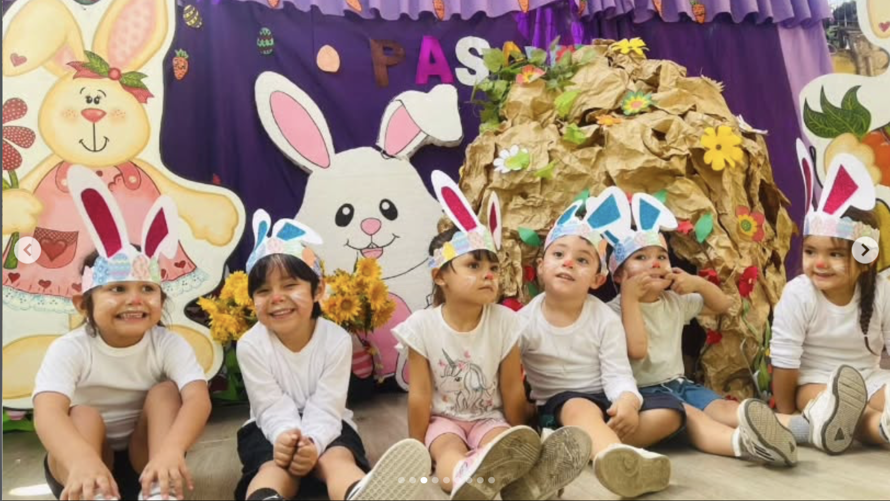 Niños en una fiesta de Pascua, usando orejas de conejo hechas con papel, sentados en el suelo con decoraciones de conejitos y flores de fondo.