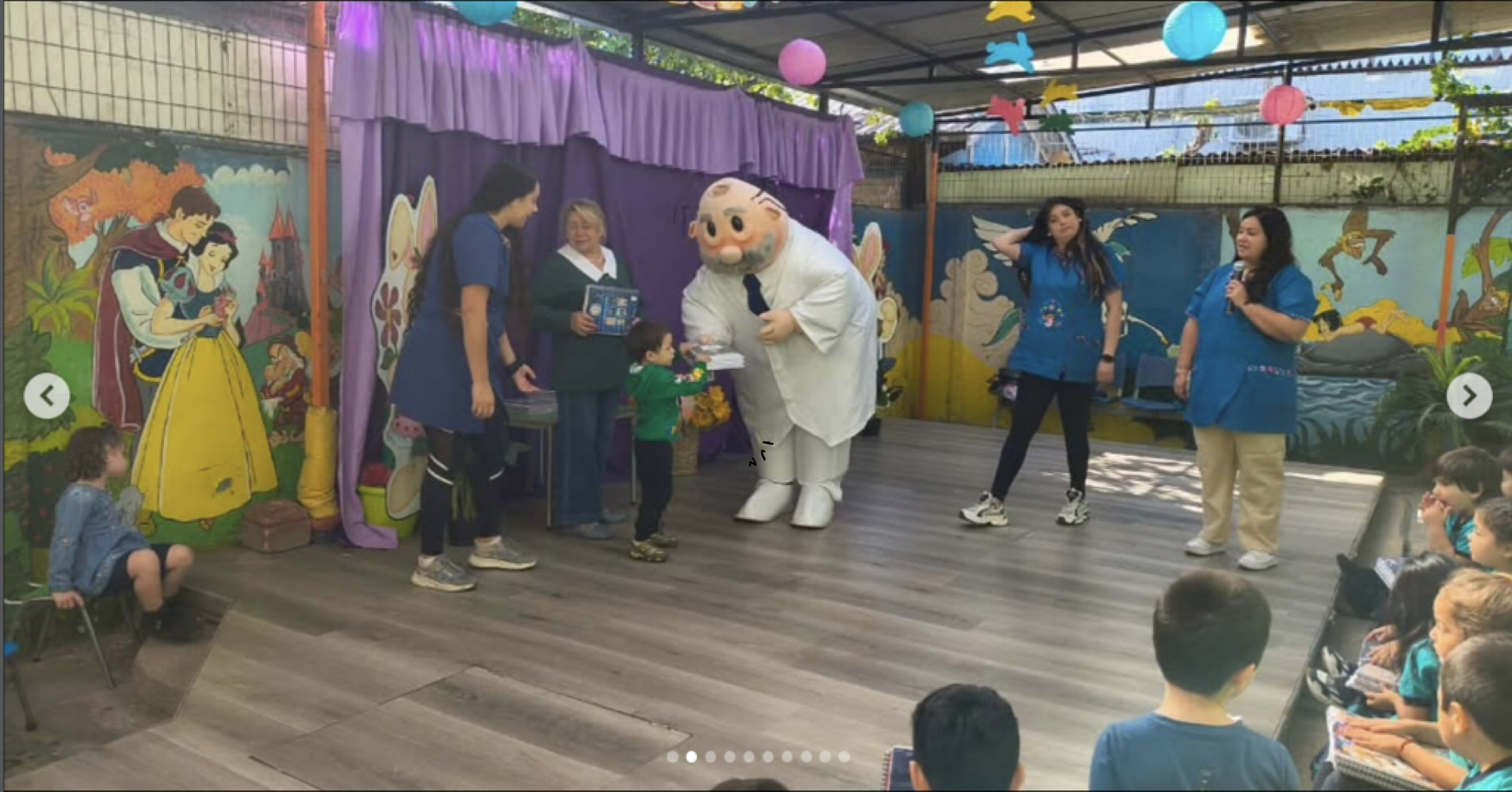 Niños y adultos participando en un show infantil con un personaje disfrazado de anciano, en un escenario decorado con personajes de cuentos y murales coloridos.