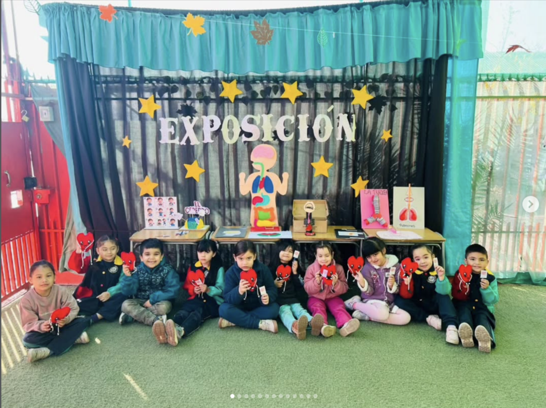 Grupo de niños en una exposición escolar sobre anatomía, sosteniendo corazones de cartón y mostrando modelos educativos en una mesa con decoraciones de hojas y estrellas en el fondo.