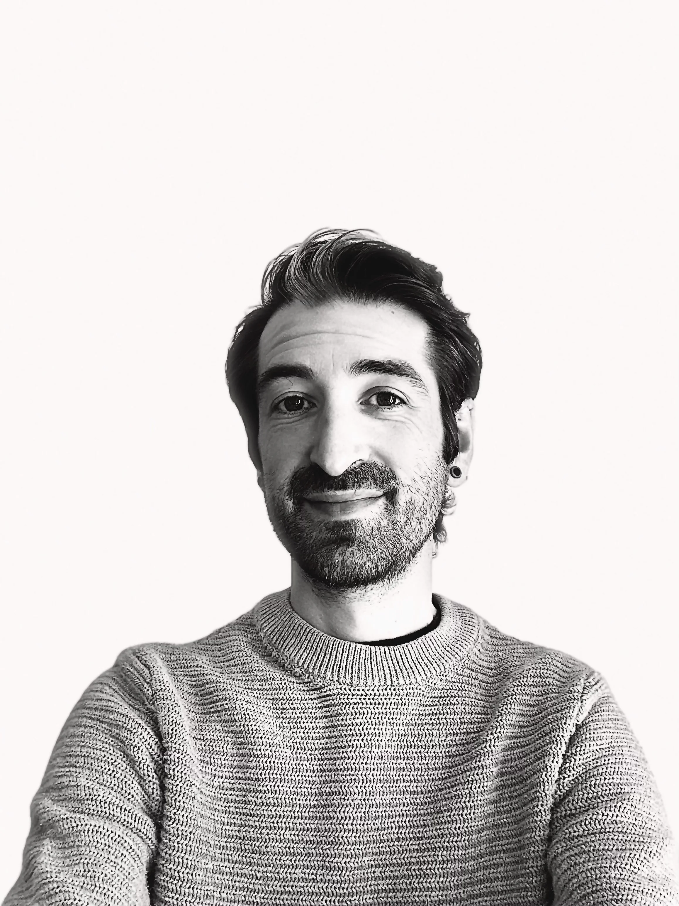 Black and white photo of a smiling man wearing a knitted sweater, with styled hair and a beard, against a plain light background.