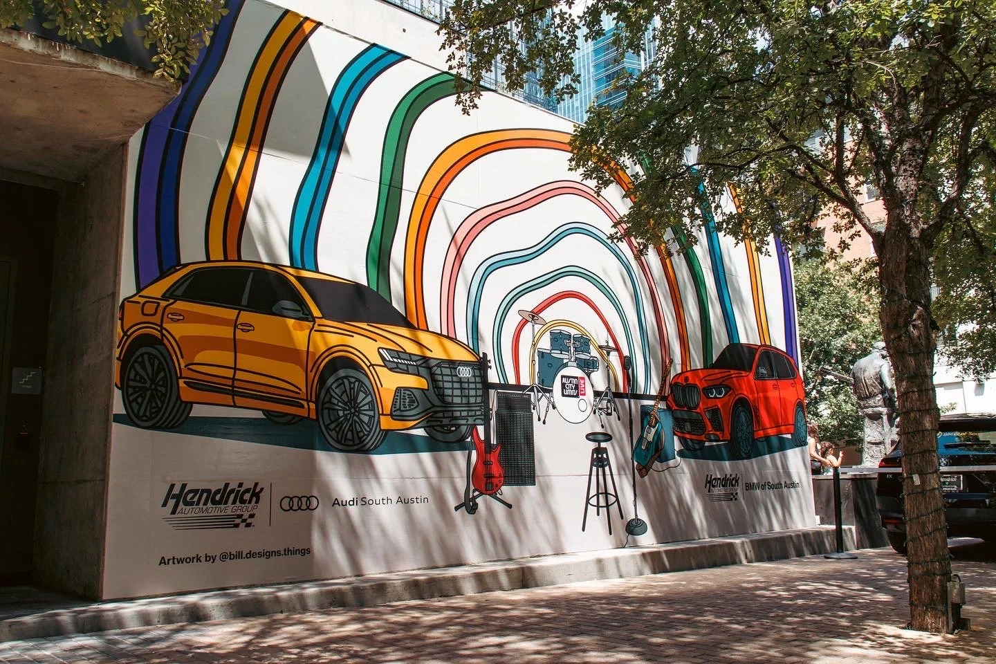 Colorful mural artwork featuring two cars, musical instruments, and rainbow-like arching lines on an outdoor wall, with trees and sidewalk in the foreground.