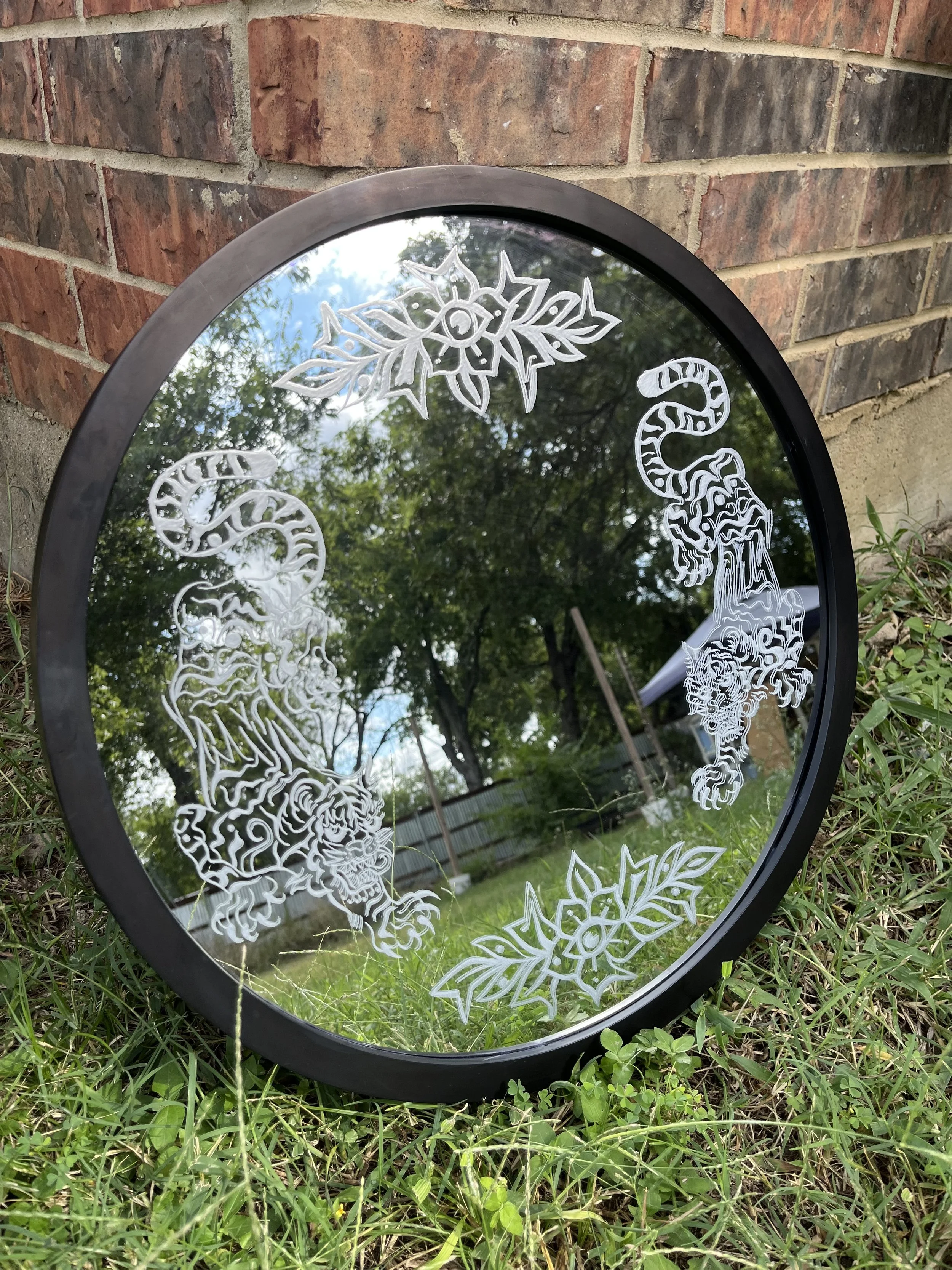 A circular mirror with Christmas-themed etchings of holly and a reindeer, placed outdoors on grass near a brick wall, reflecting trees and the sky.