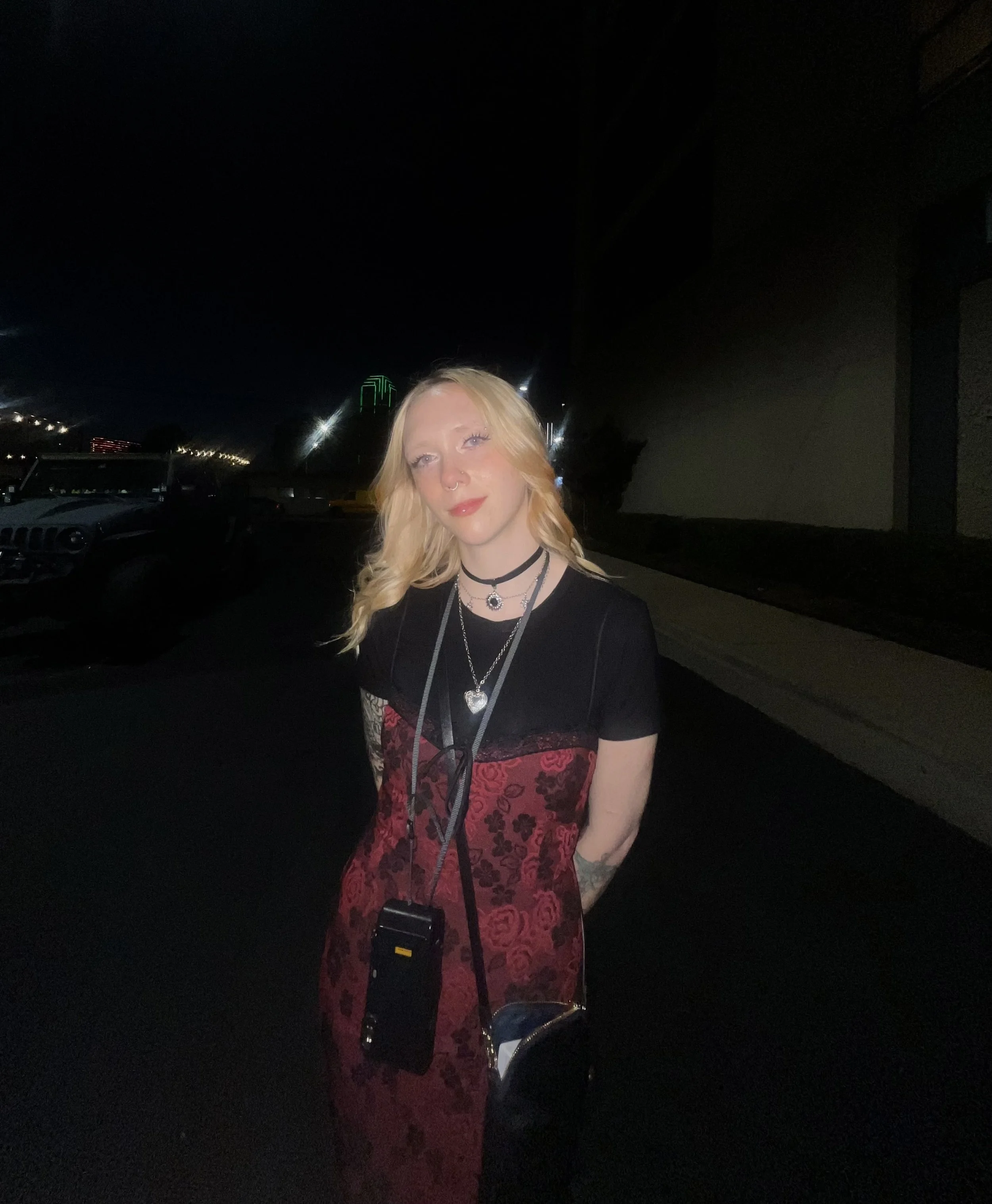 A person with long blonde hair, wearing a red and black floral dress, multiple necklaces, and a septum piercing, standing outdoors at night in a parking lot with cars and city lights in the background.