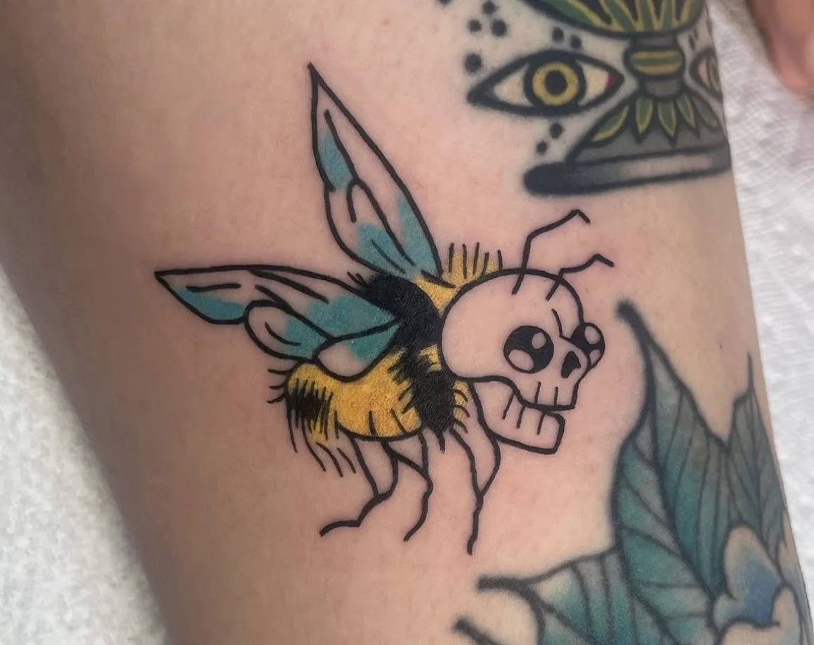 Tattoo of a cartoon-style bee with a skull face and butterfly wings.