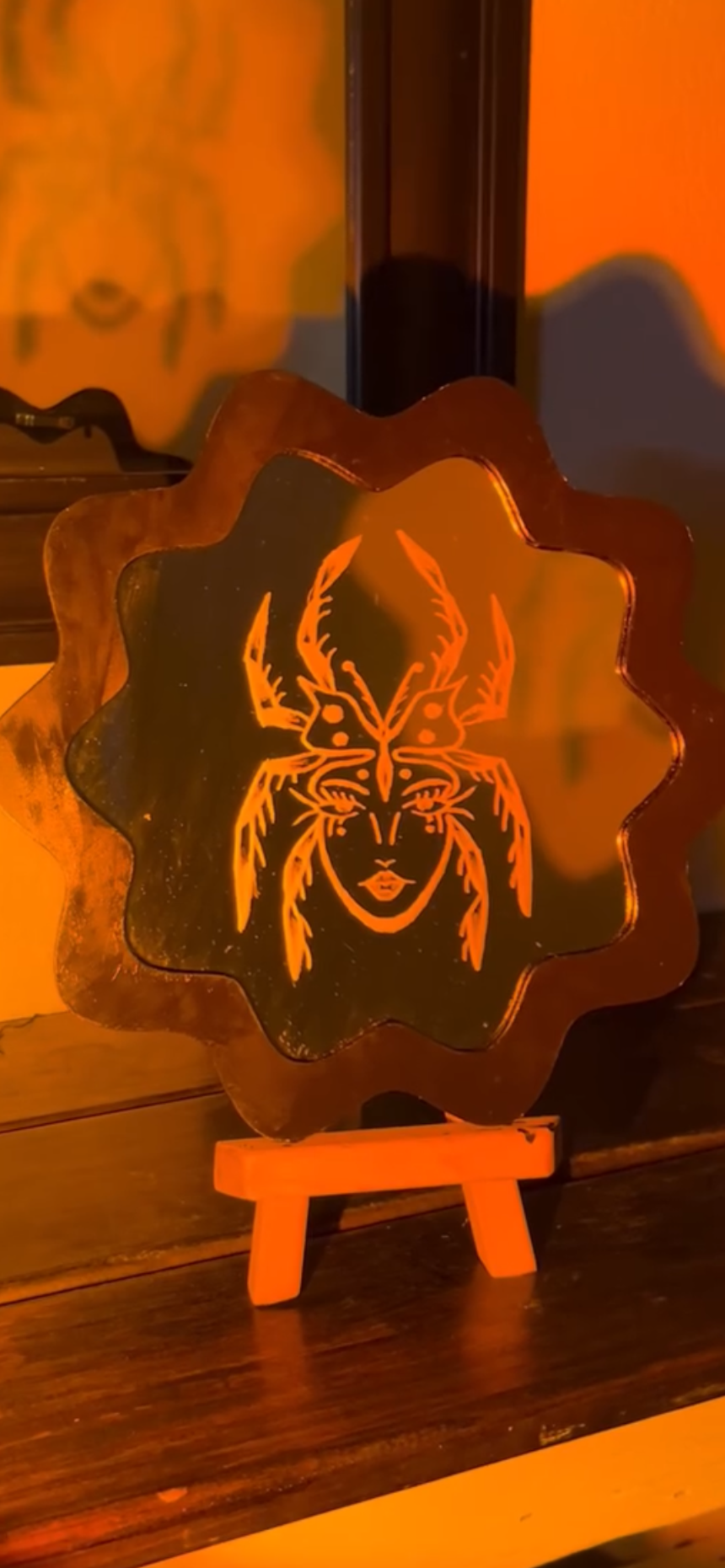 Illuminated decorative lamp featuring a design of a woman's face with antlers and a butterfly on the forehead, placed on a small wooden easel in a dimly lit room.