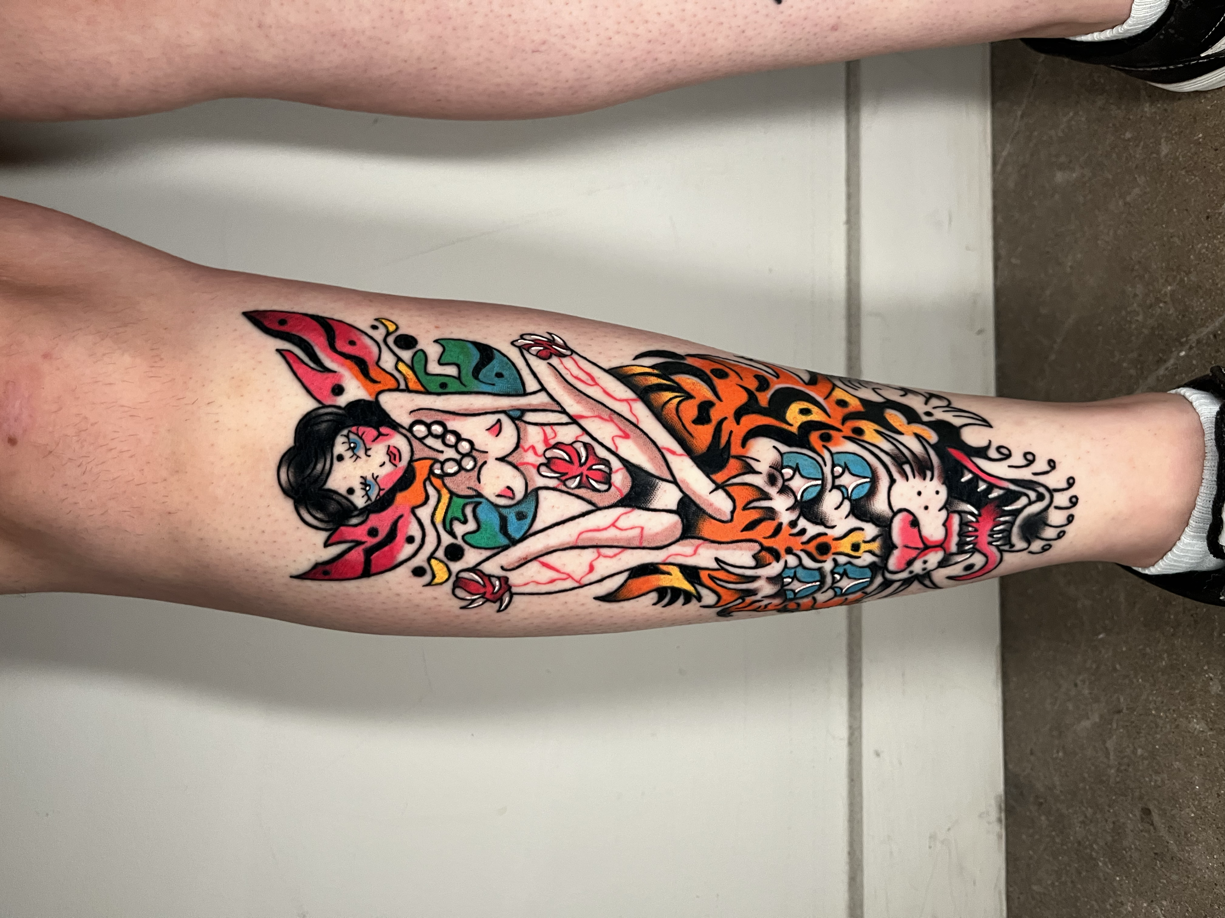 A colorful tattoo on a person's leg featuring a fierce tiger with an open mouth, sharp teeth, and prominent whiskers, surrounded by flames and abstract patterns.