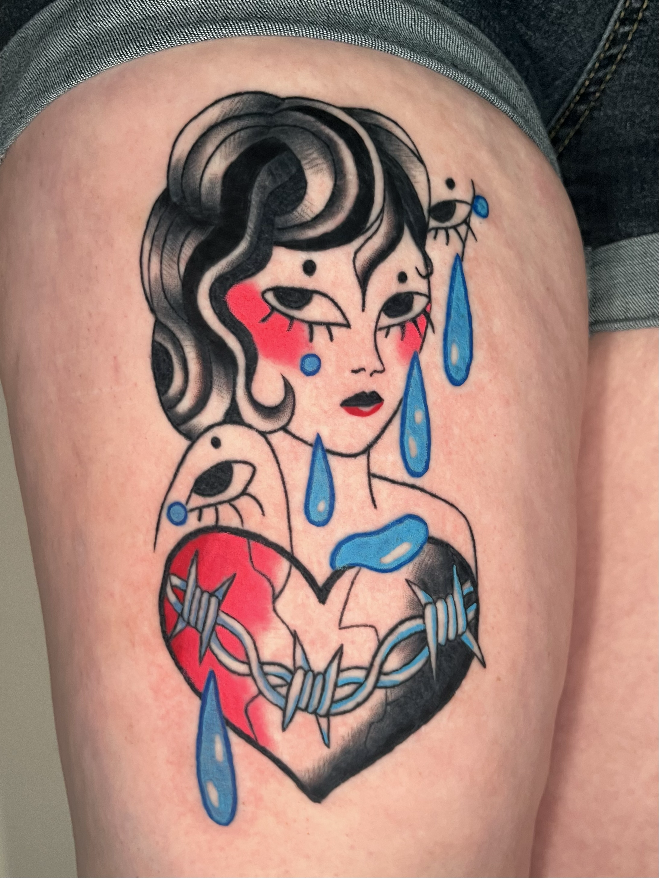 Tattoo of a woman with black hair, red cheeks, and tears, surrounded by a heart and with blue tears and drops.