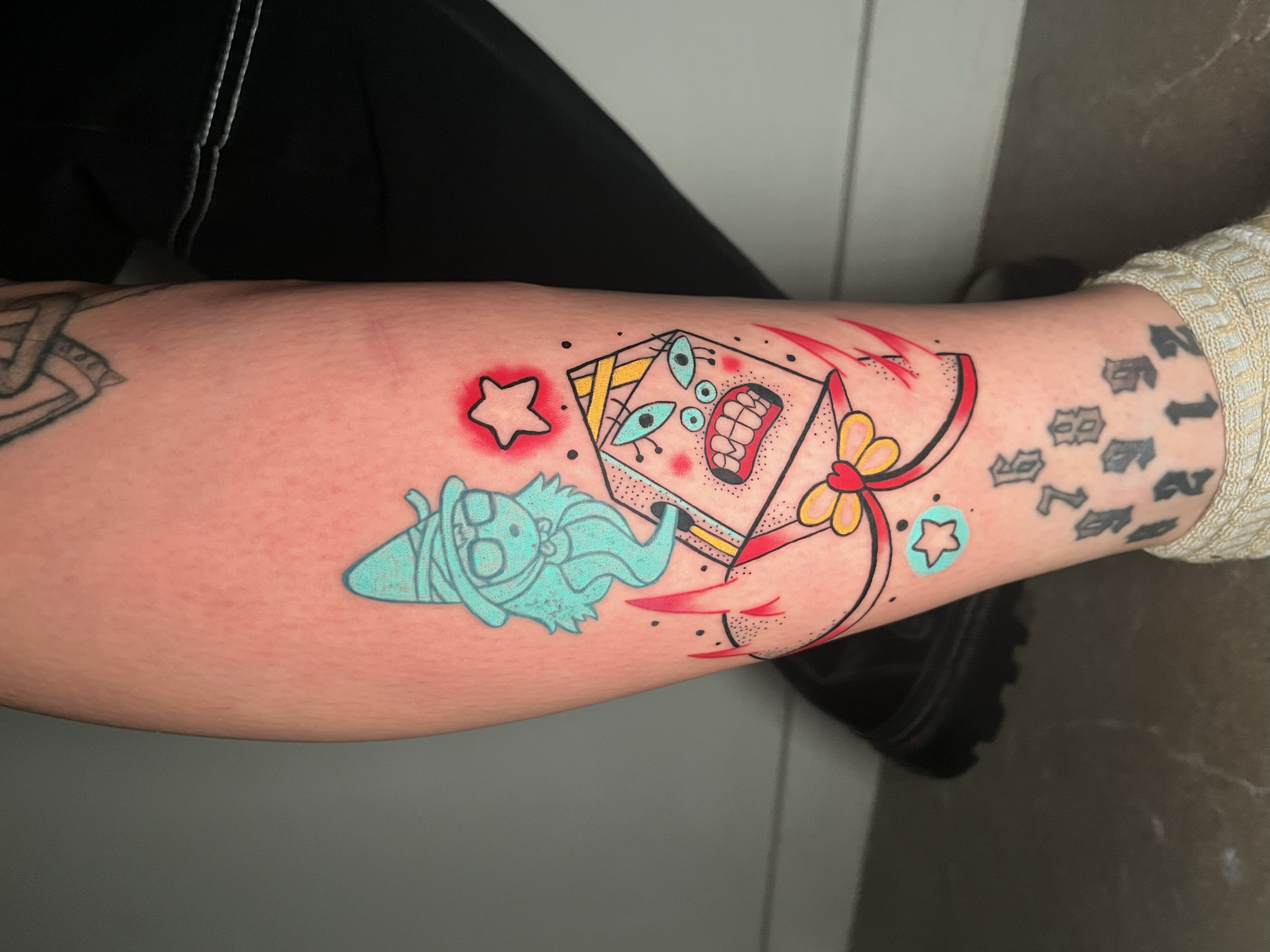 Tattooed person's lower arm with colorful tattoo design including a cartoon girl with glasses and wizard hat, a star with red glow, a yellow and pink bow, a doomsday device, and numbers.