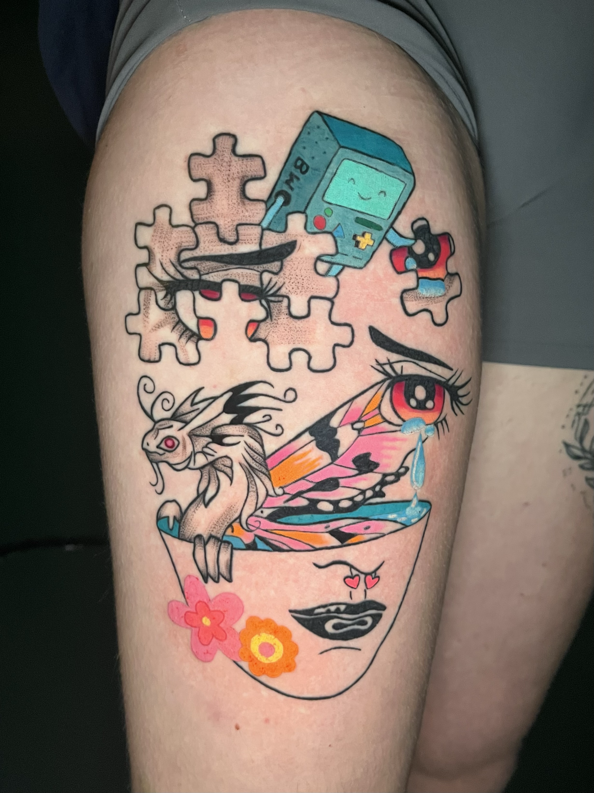 Tattoo of a smiling vintage Game Boy with a cord, puzzle pieces, a dragon, a face with multicolored flowers, and hearts.