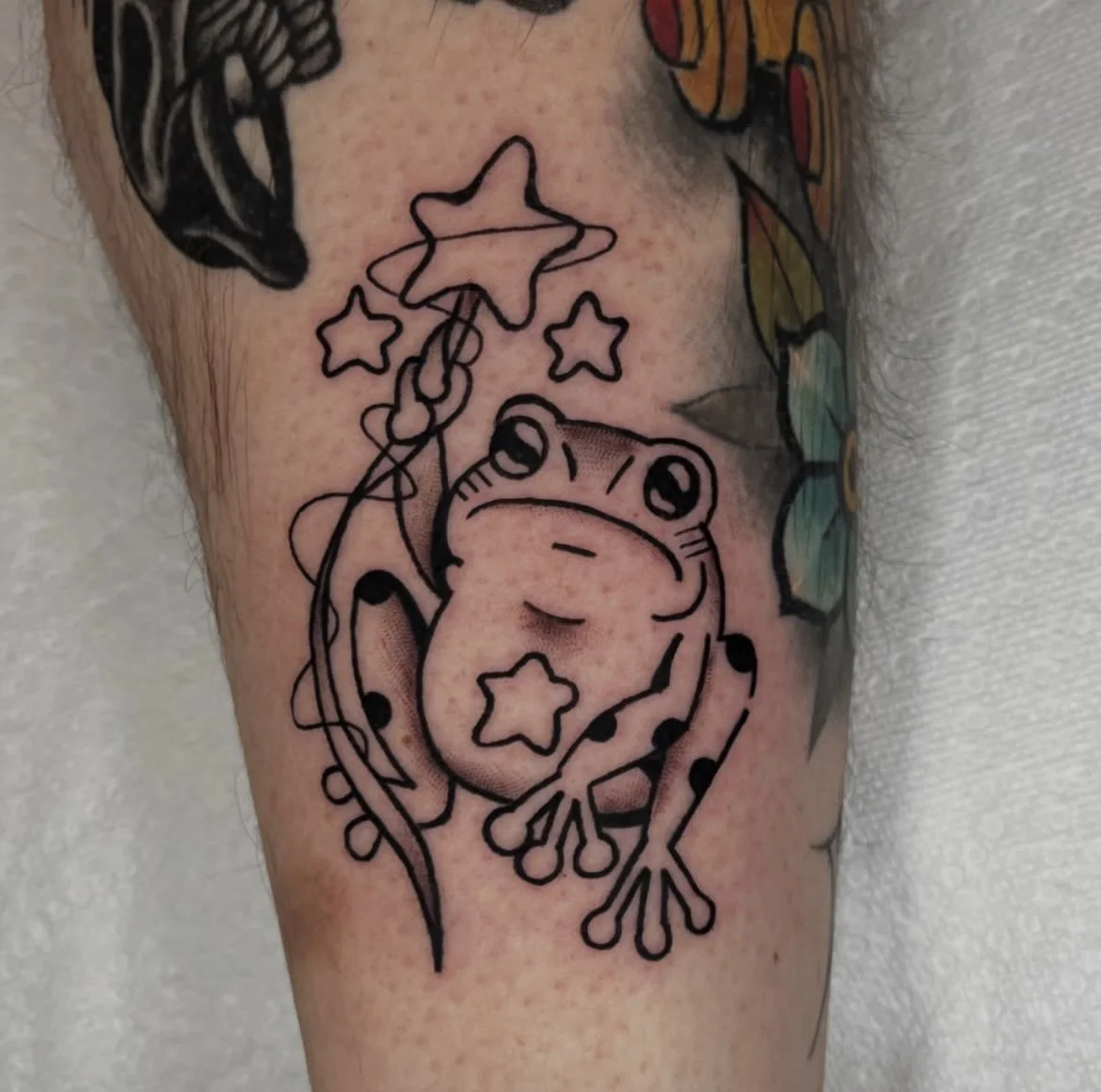 Tattoo of a cartoon frog holding a star-shaped balloon, with additional star-shaped details.