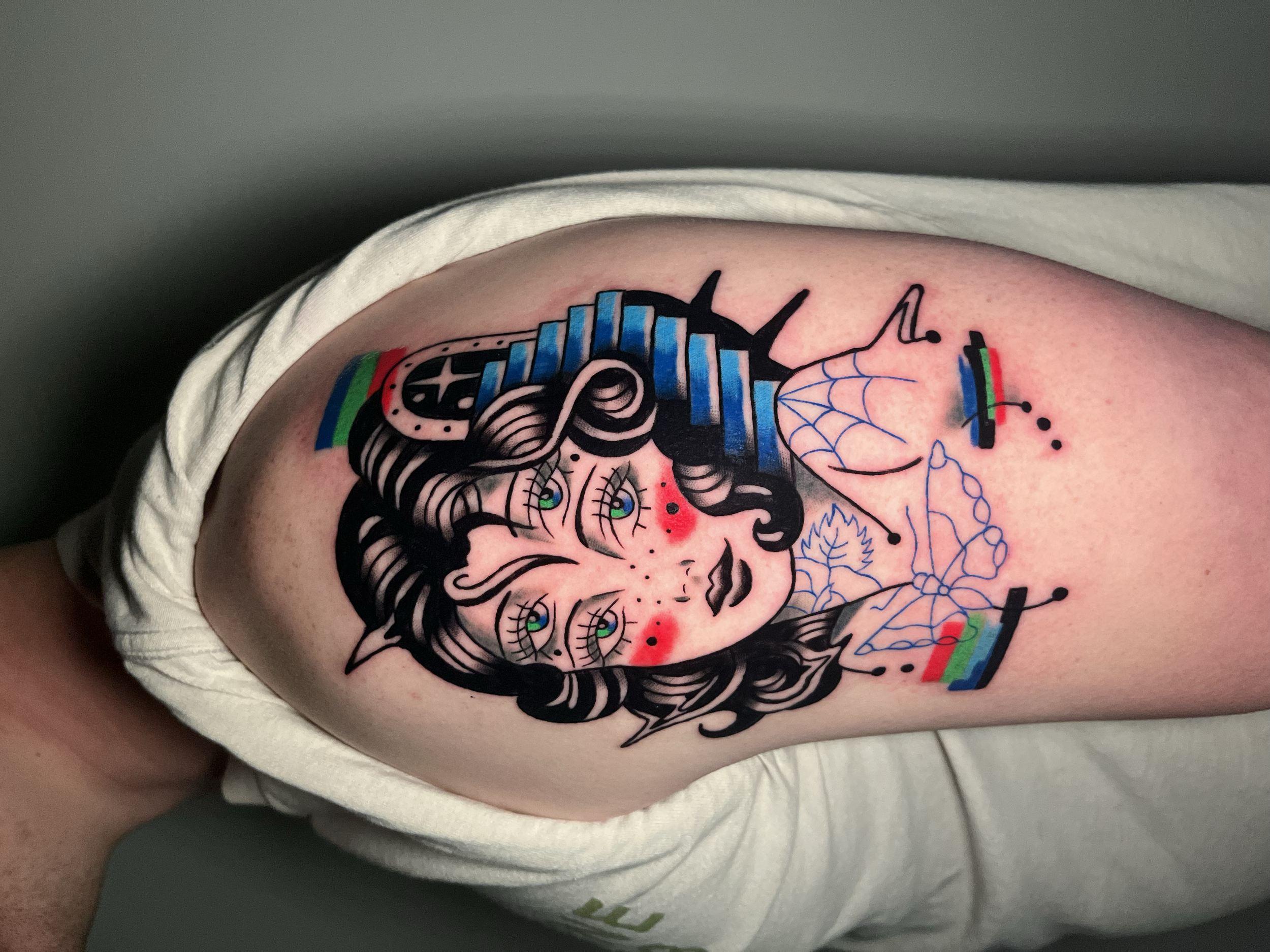 Tattoo of a woman with multi-eyed face, surrounded by colorful elements, on upper arm.