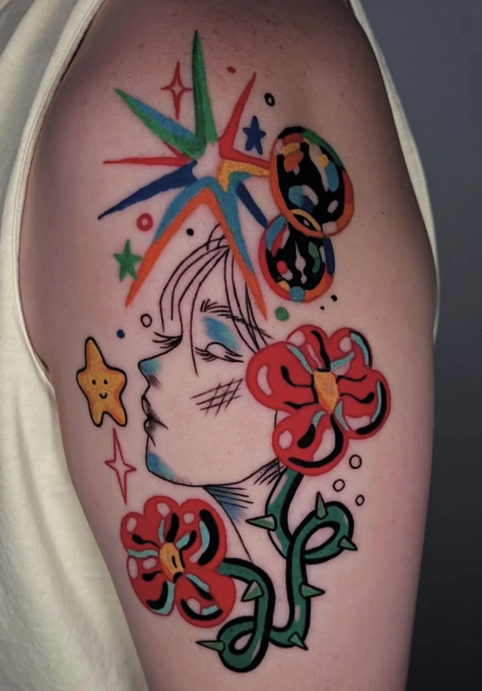 Colorful tattoo of a woman's face with closed eyes, surrounded by a rainbow-colored starburst, a butterfly, red flowers with black accents, a small yellow star, and various small stars and circles.