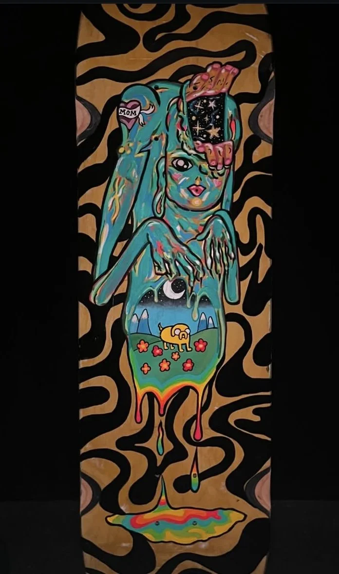Colorful surreal artwork featuring a woman with long blue hair, a tattoo that says 'Mom,' and a hand covering part of her face. The woman’s body depicts a landscape with a yellow dog, flowers, mountains, and a starry sky, with rainbow-colored drips and abstract black shapes on a wooden background.