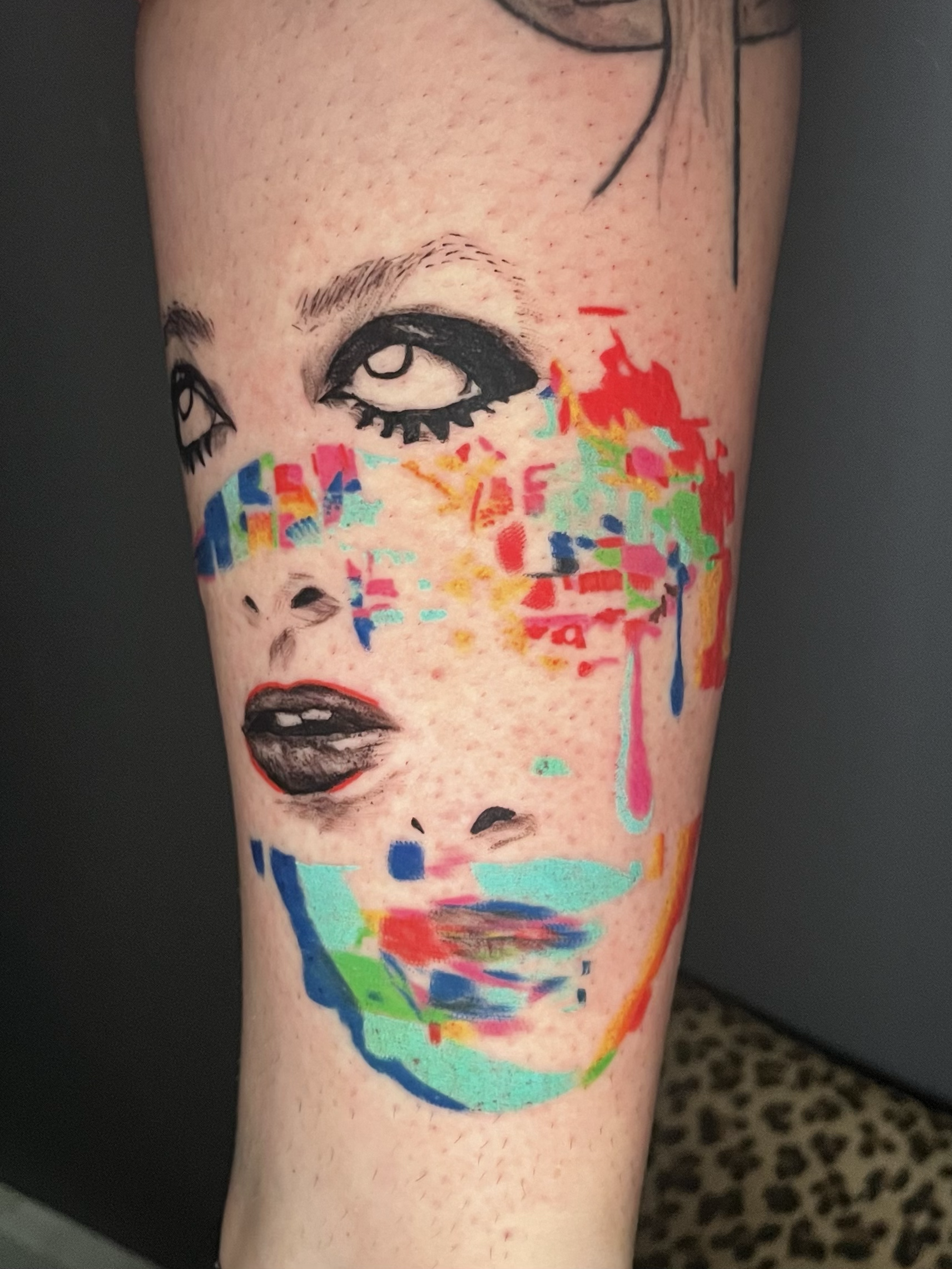 Tattoo of a woman's face with makeup, with colorful abstract paint splashes around it on a person's arm.