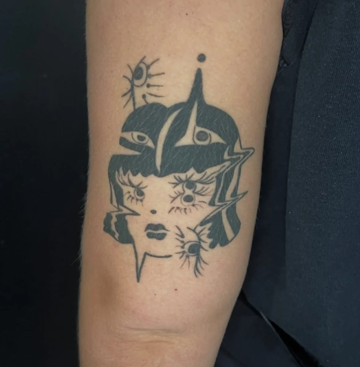 Black tattoo of a stylized woman's face with multiple eyes, eyelashes, and lips, surrounded by abstract shapes, lightning bolts, and small eye symbols.