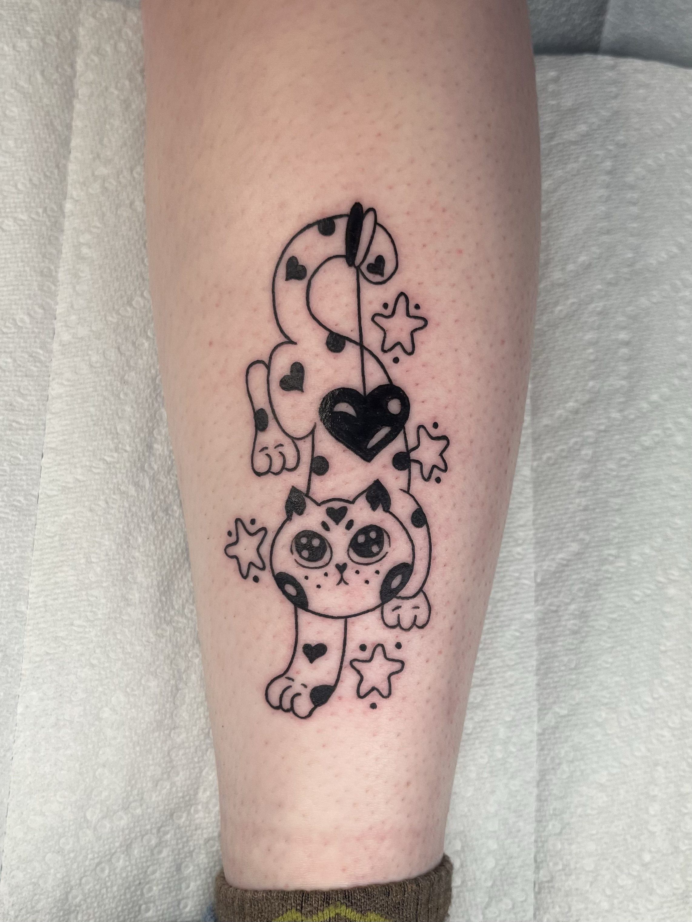 Tattoo on a person's leg featuring a cartoonish unicorn with big eyes, surrounded by stars, hearts, and small decorative elements.