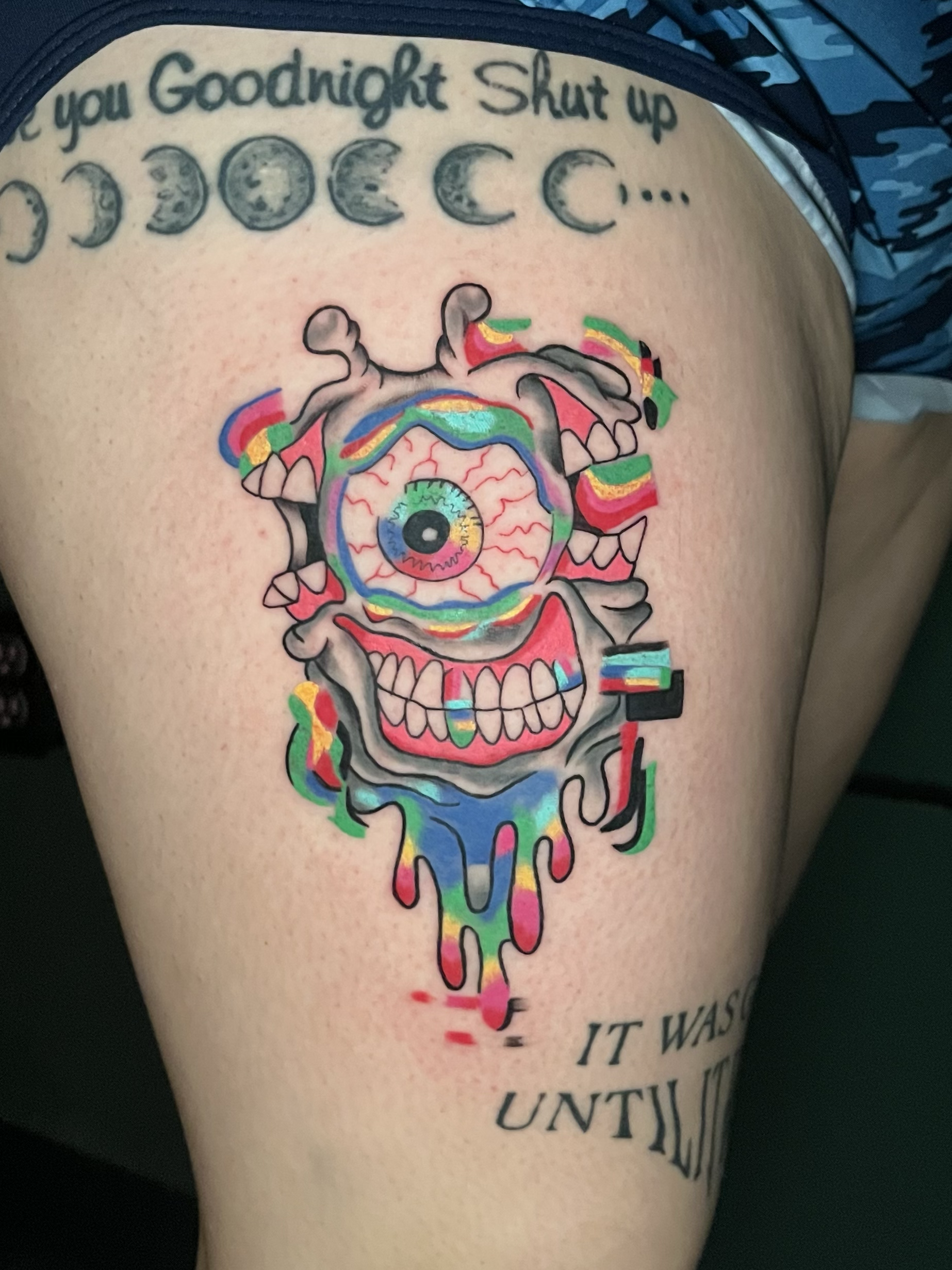 Colorful tattoo of a melting skull with an iris in the center, surrounded by partial text including "Goodnight" and "Shut up."