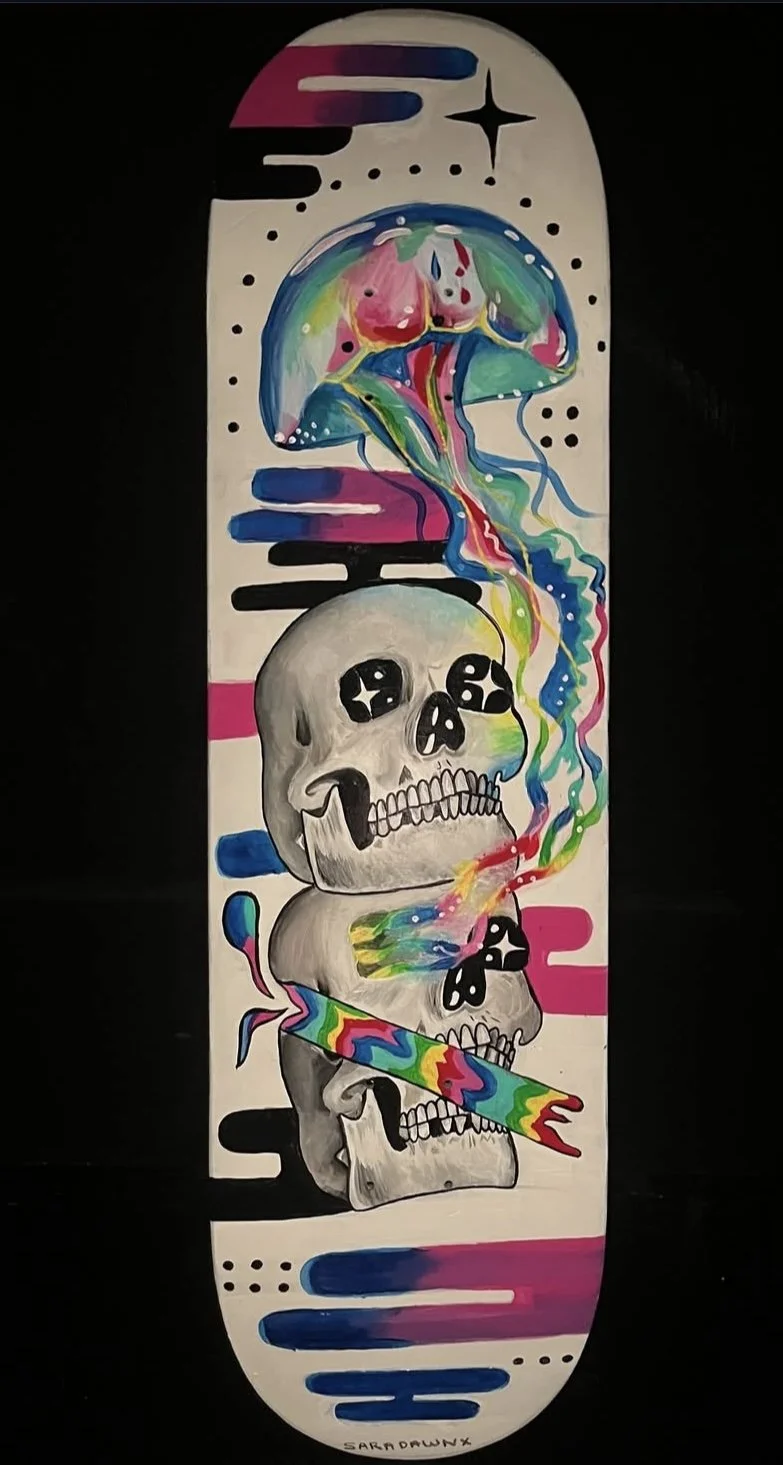 A skateboard deck features a colorful, surreal painting of stacked skulls with expressive, starry eyes and rainbow smoke or energy emanating from the top, with abstract black, pink, and blue lines and dots in the background.