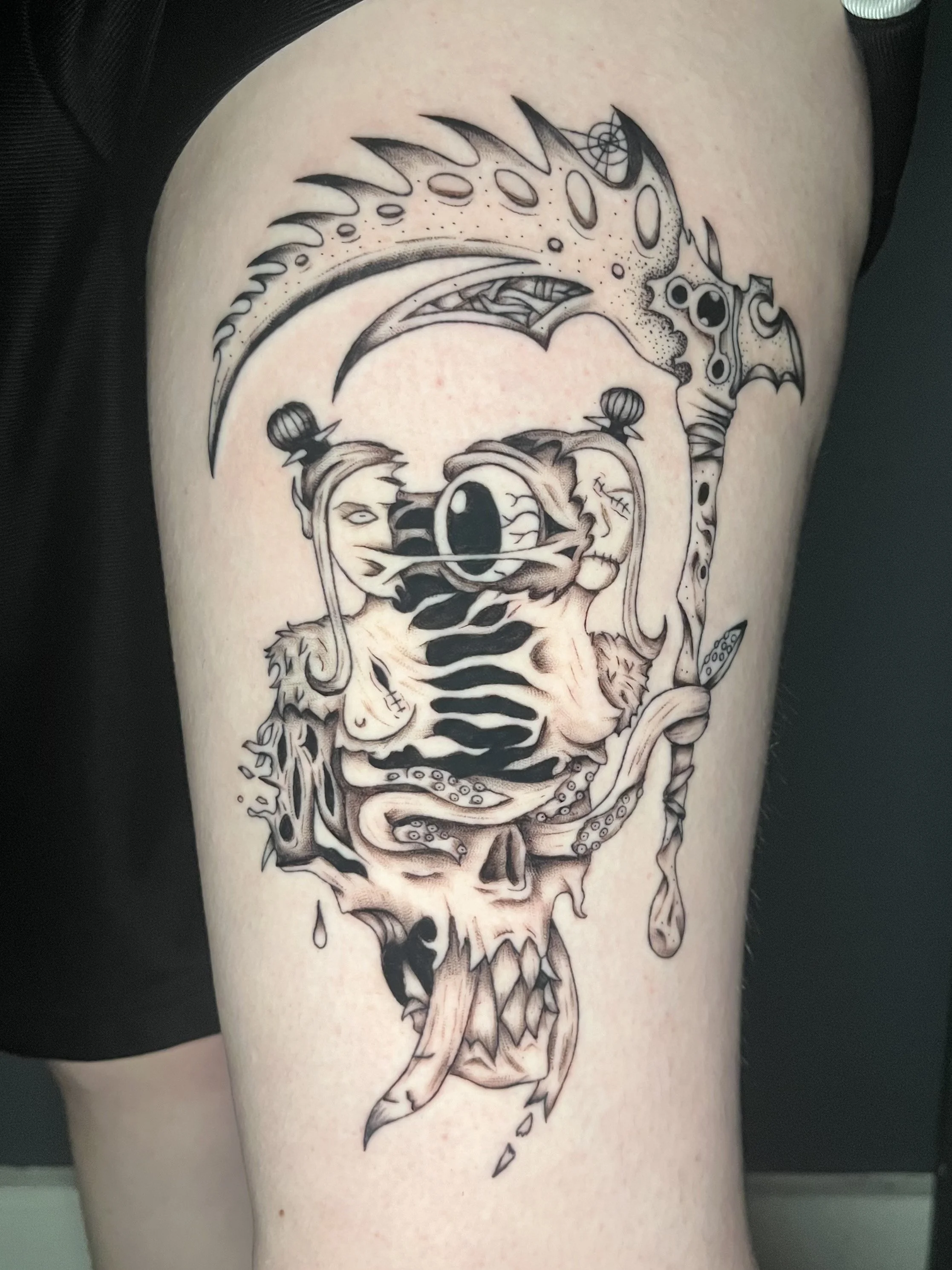 A black and gray tattoo on a person's thigh featuring a skull with a snake emerging from its eye socket, with a dragonfly perched on top of the skull, and a large curved sword behind the skull.