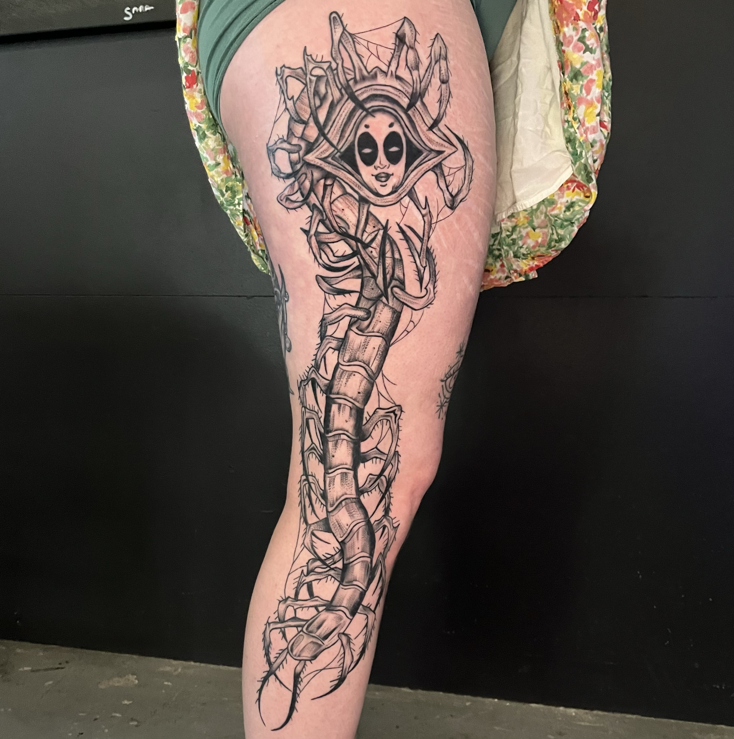 Tattoo of a cartoonish alien face with large black eyes, surrounded by spider webs and a skeleton, on a person's thigh.