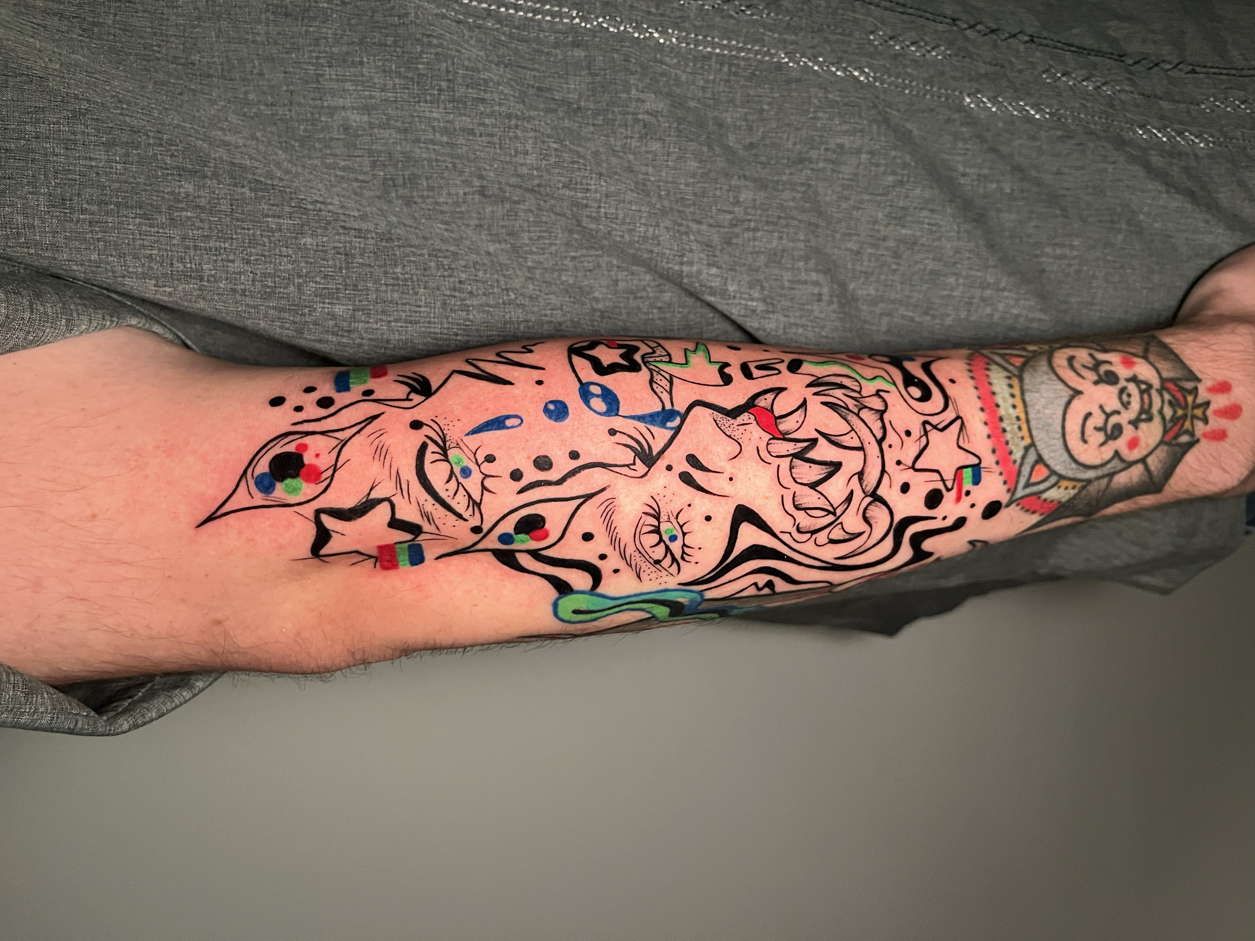 Colorful tattoo sleeve with various cartoonish characters and abstract details on a person's arm, with part of clothing visible.