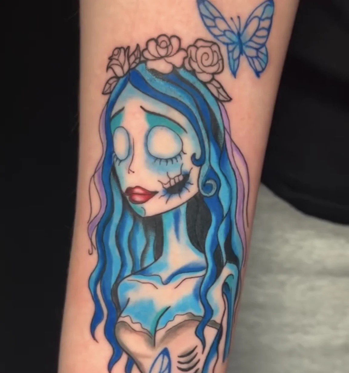 Tattoo of a woman with blue hair and a death-themed face, flowers on her head, and a butterfly nearby.