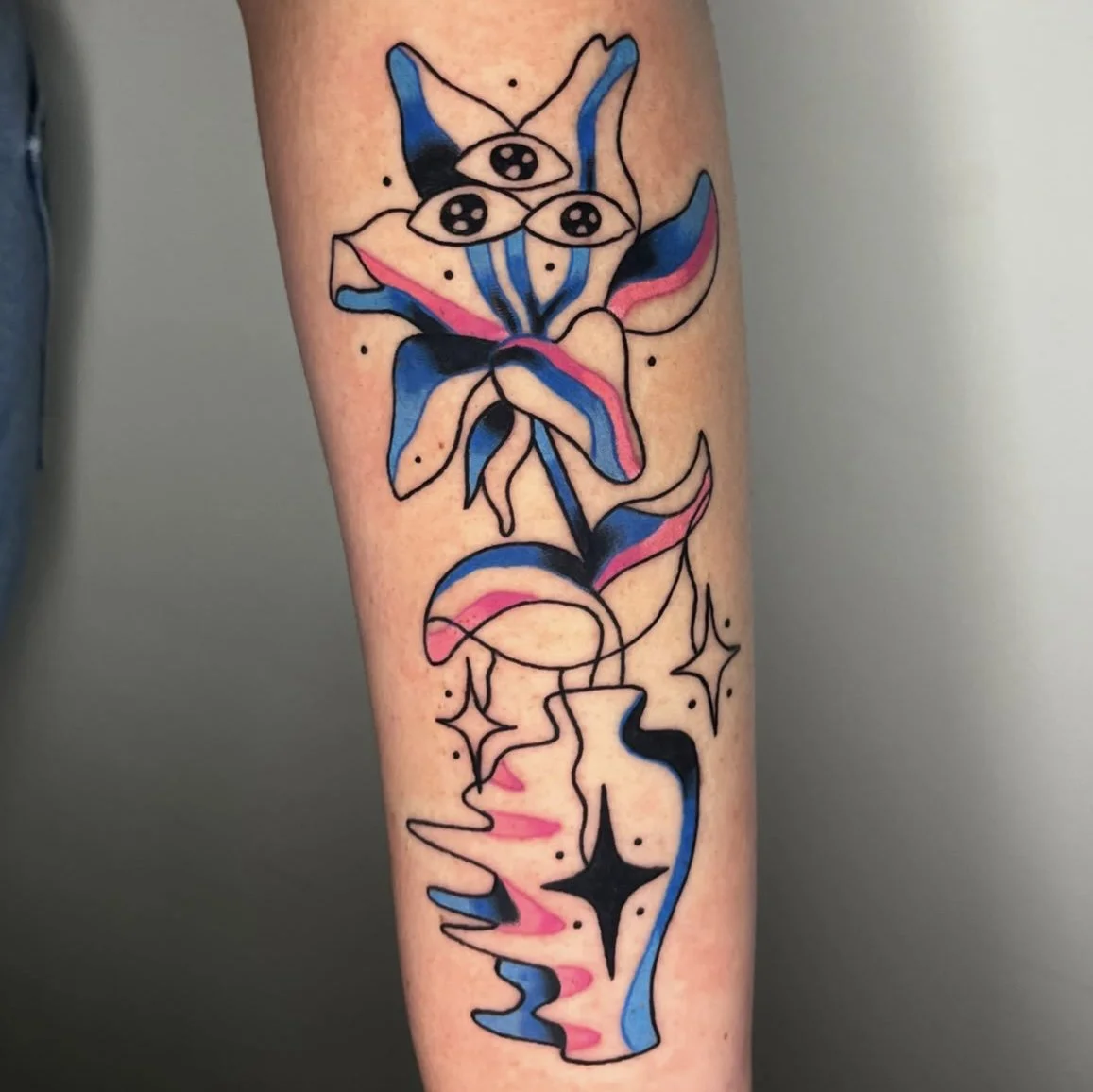Colorful abstract tattoo of a flower with three eyes at the top, a stem, and a vase at the bottom, featuring blue, pink, black, and white colors.