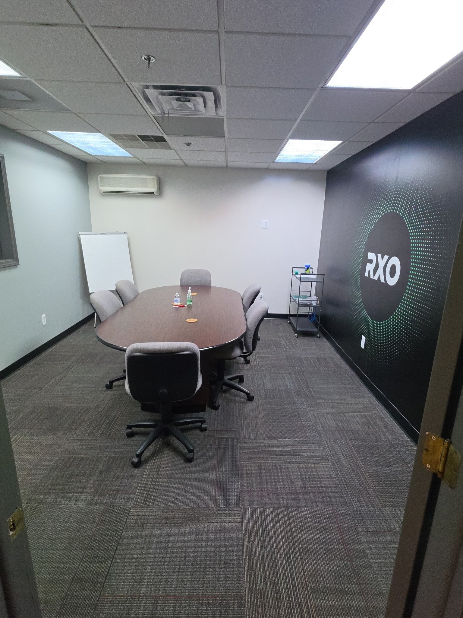 An empty conference room with a large oval table, six chairs, a whiteboard, a small cart, water bottles, and a large backdrop with 'RXO' written on it.