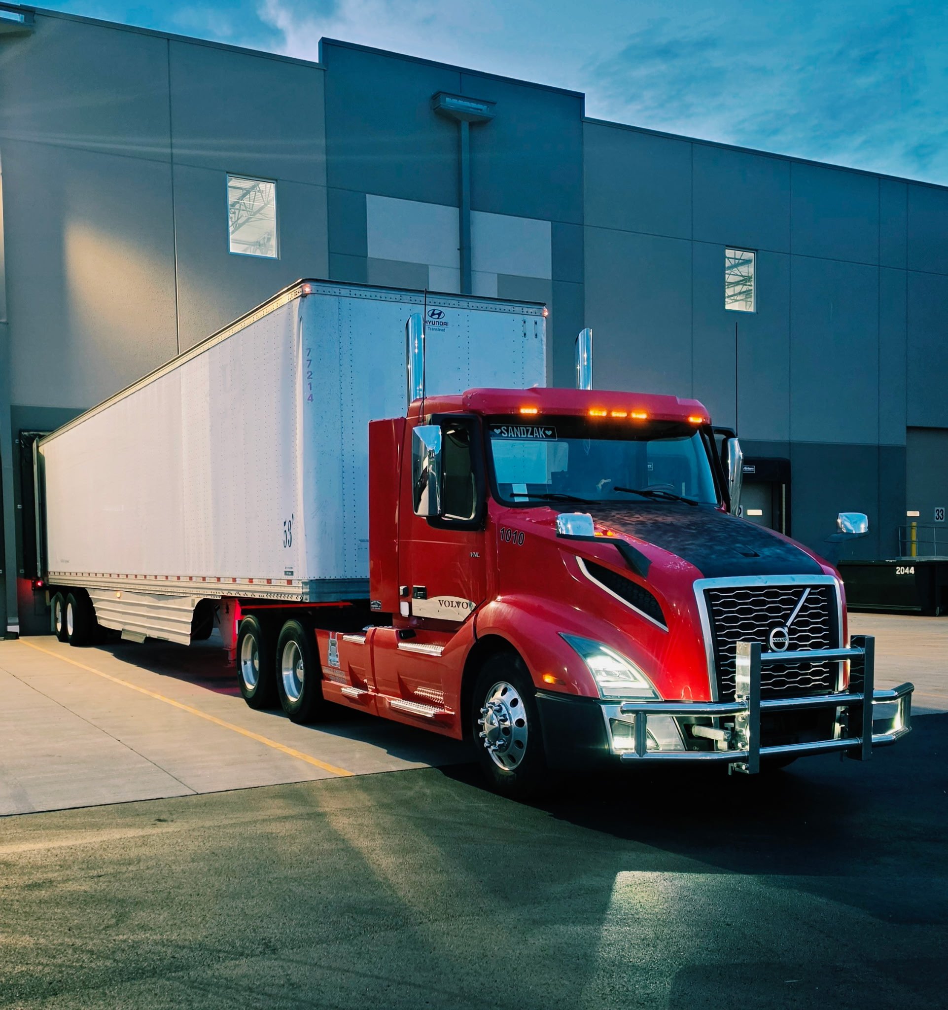 Red semi-truck with box trailer parked in a lot with gray pavement. Building with dark gray walls and small windows in the background. Clear sky with a few clouds overhead.