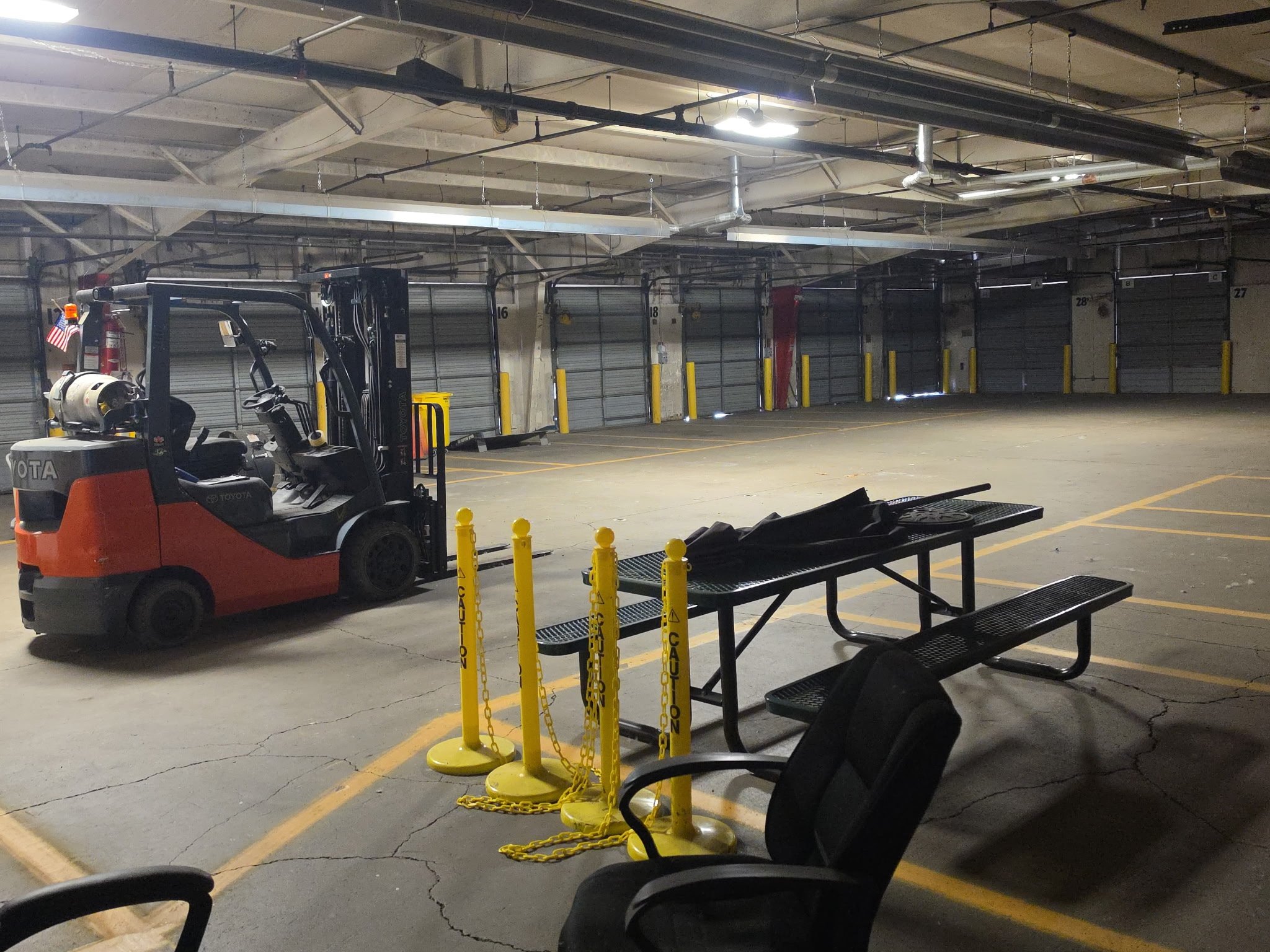 Inside an empty garage or storage unit with closed metal doors, a forklift is parked on the left side, and yellow caution chains are in the foreground. The ceiling shows exposed pipes and lighting.