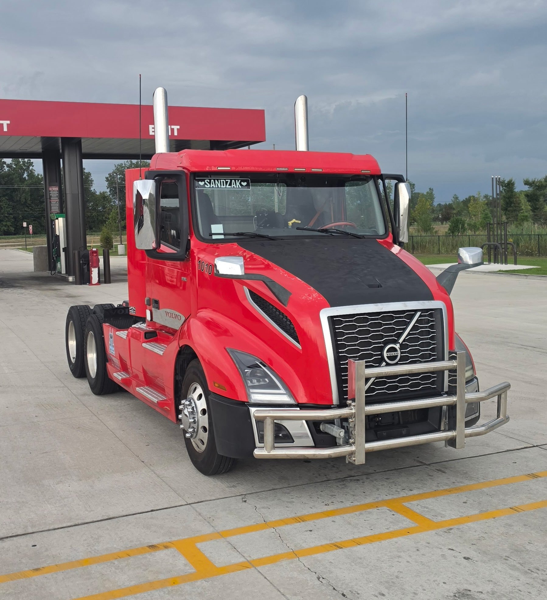 Red semi-truck parked at a gas station, with the name 'SANDZAK' on the front windshield. The truck is without a trailer and is positioned near the fuel pumps on a concrete lot with yellow parking lines. The sky is overcast with dark clouds.