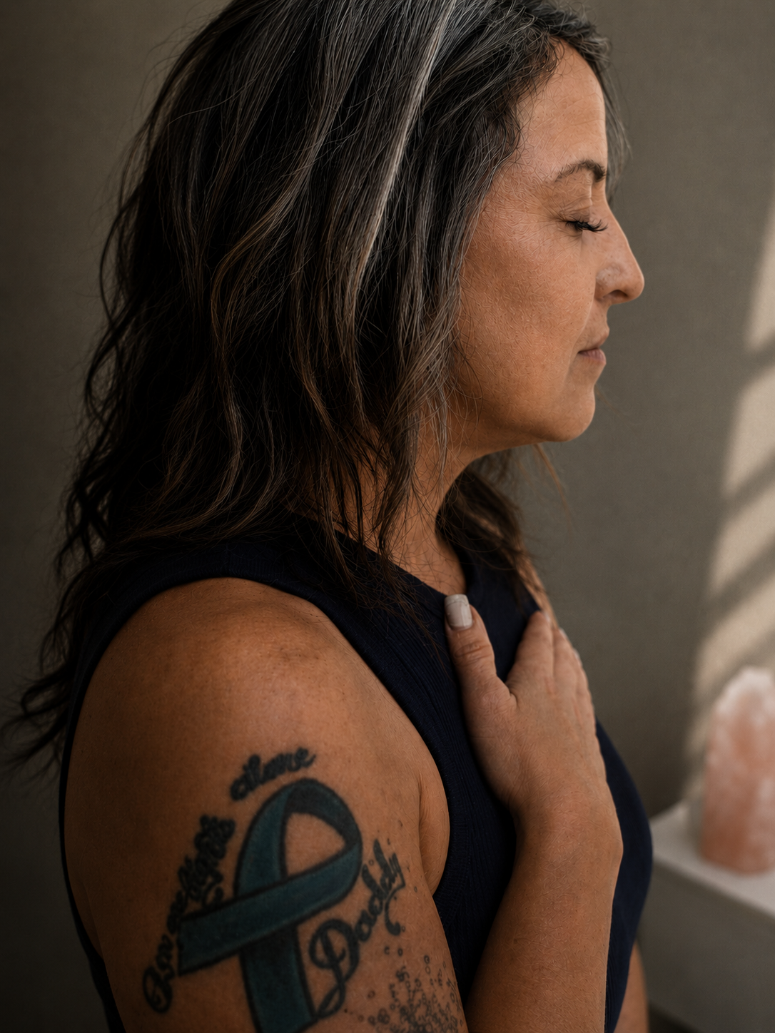 Side profile of a woman with gray hair and a tattoo on her upper arm, standing with her hand on her chest, eyes closed, indoors.
