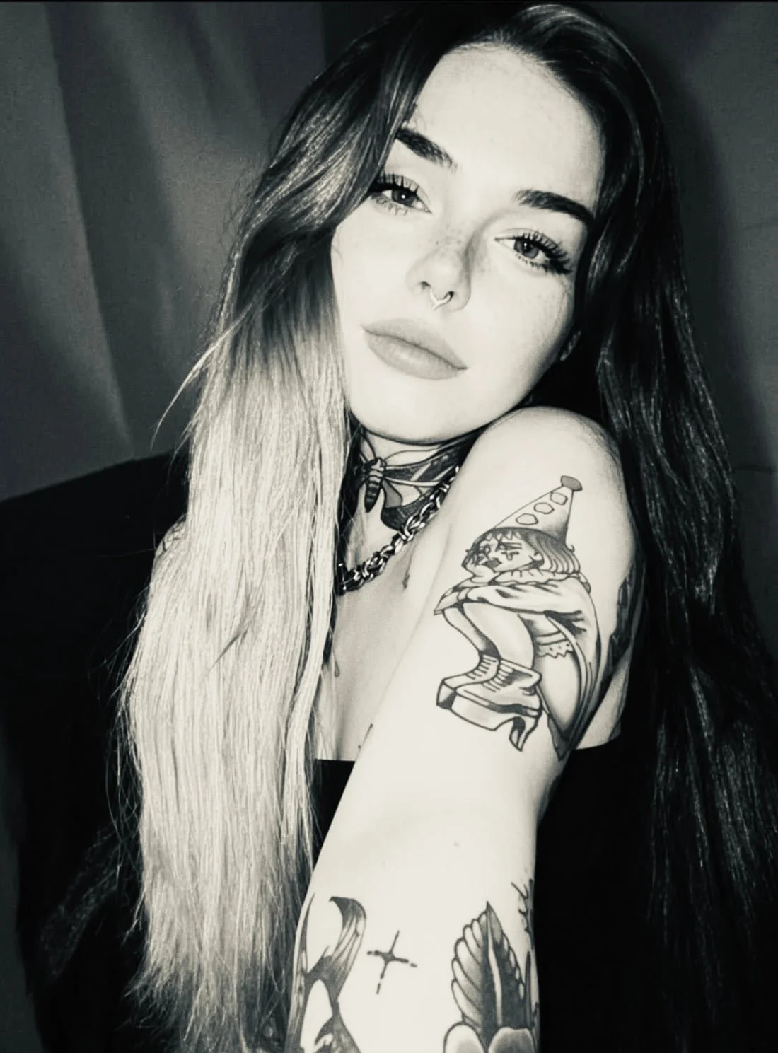 A black and white photo of a young woman with long dark and blonde hair, tattoos on her arm, wearing a nose ring, necklaces, and a dark top, posing indoors with a neutral expression.