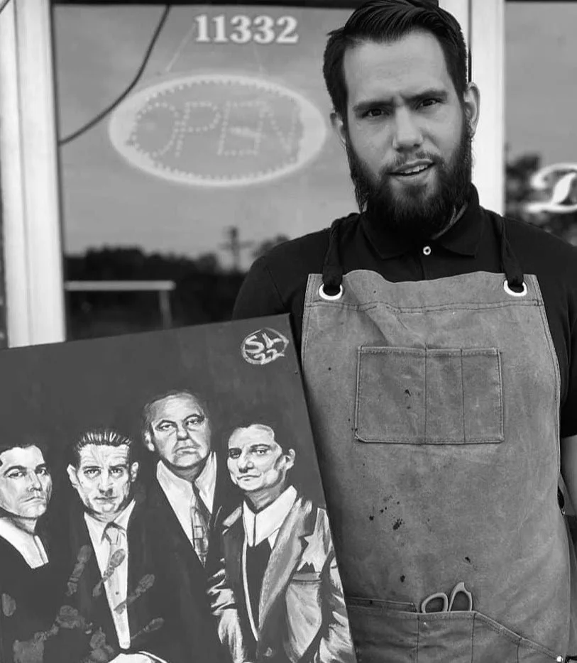 A man with a beard wearing a black shirt and apron standing outside a store, holding a painting of four men. There is an 'OPEN' sign in the window behind him.