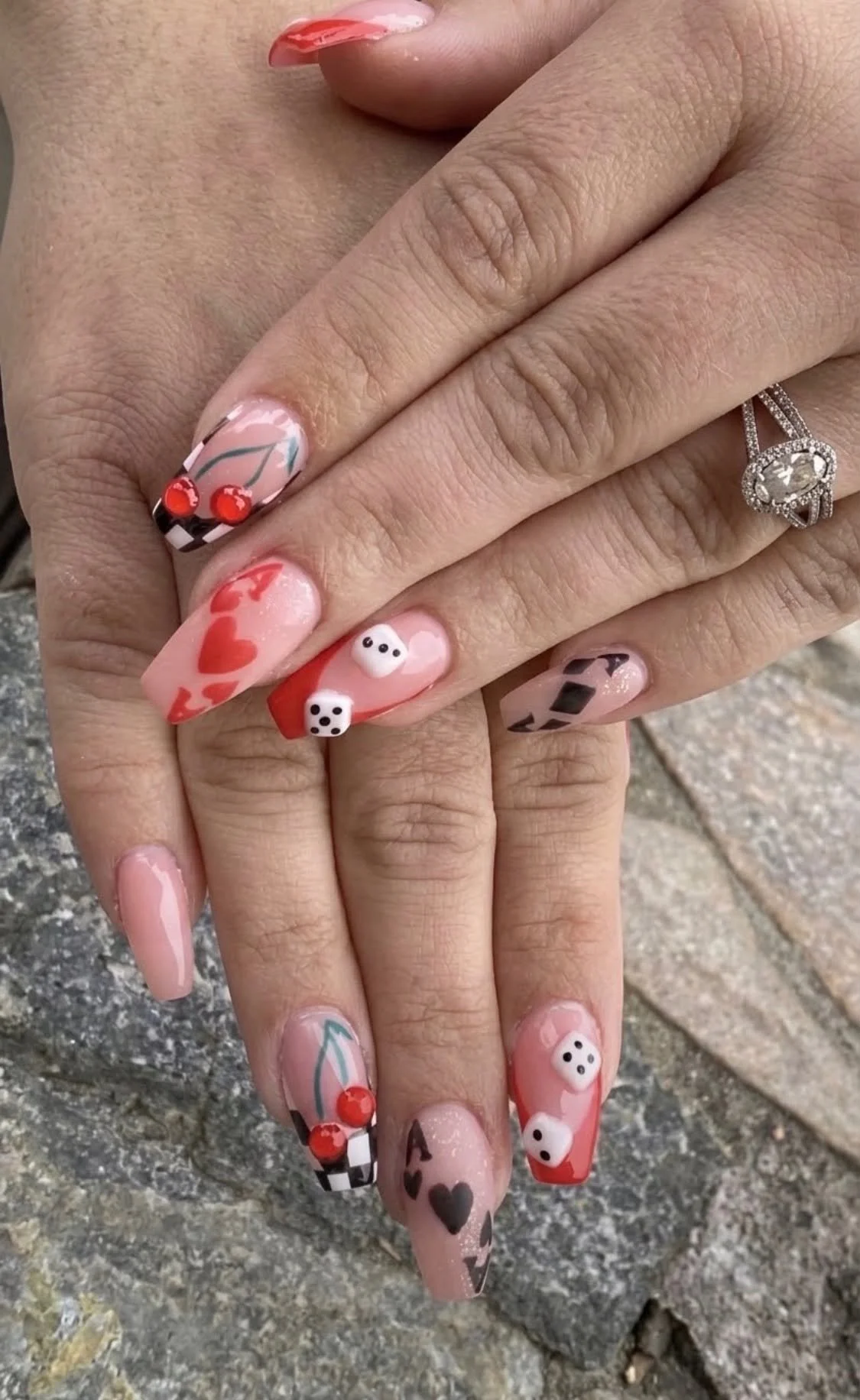 Close-up of hands with decorated nails featuring hearts, dice, cherries, and a small ghost, wearing a wedding ring with a large central stone.