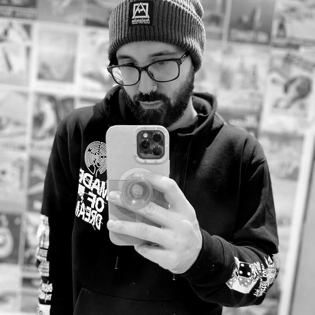 A man taking a selfie in a mirror, wearing glasses, a beanie, and a hoodie, with a background of various posters or pictures on a wall.