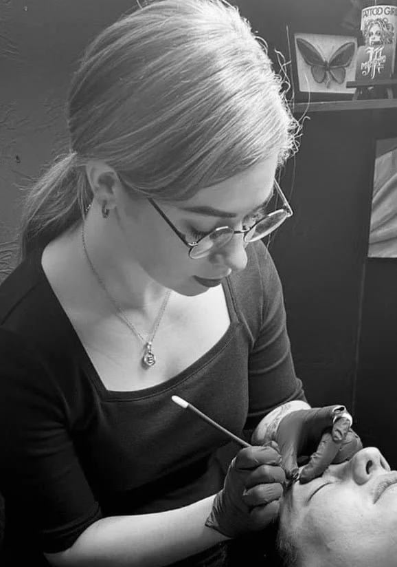 A woman with glasses and blonde hair in a ponytail is performing a cosmetic tattooing or microblading procedure on a person's eyebrows in a tattoo studio.