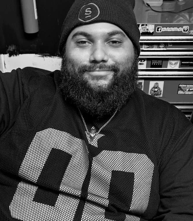 A smiling man with a beard wearing a beanie and sports jersey, sitting in front of a shelf with various gaming and media items.