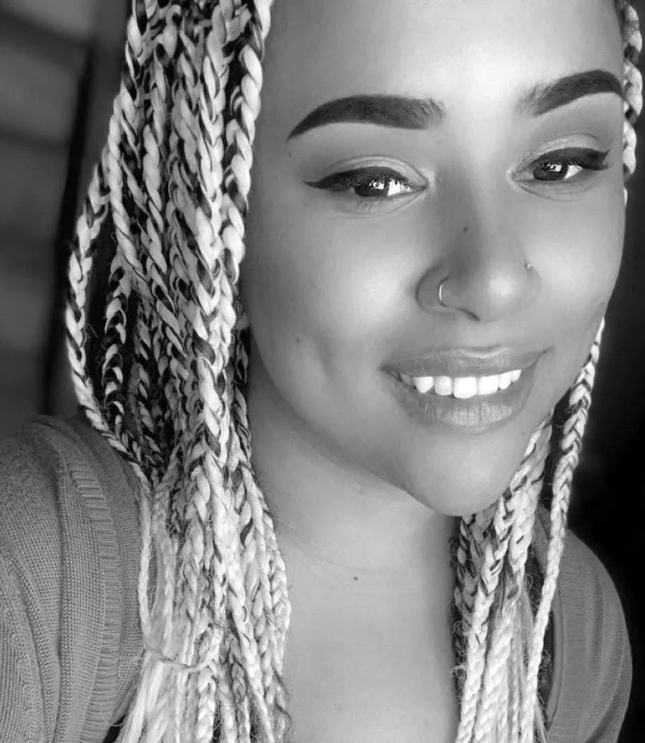 A woman with braided hair, winged eyeliner, a small nose ring, and a bright smile showing her teeth. She appears to be wearing a top with a shoulder strap.