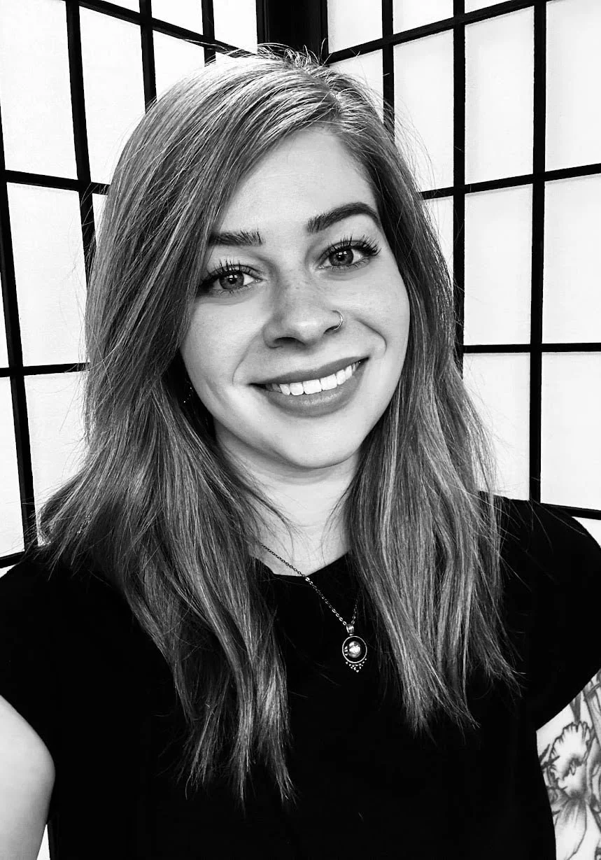 A young woman with long hair, smiling, wearing a nose ring, necklace, and a black top, standing in front of a grid-patterned background.