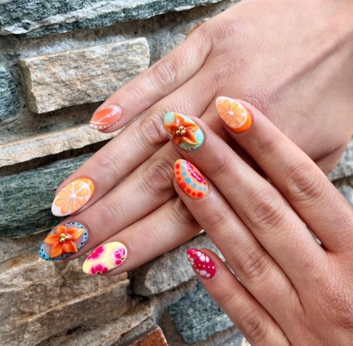 Close-up of hands with colorful, floral and polka dot nail art, positioned against a stone wall background.