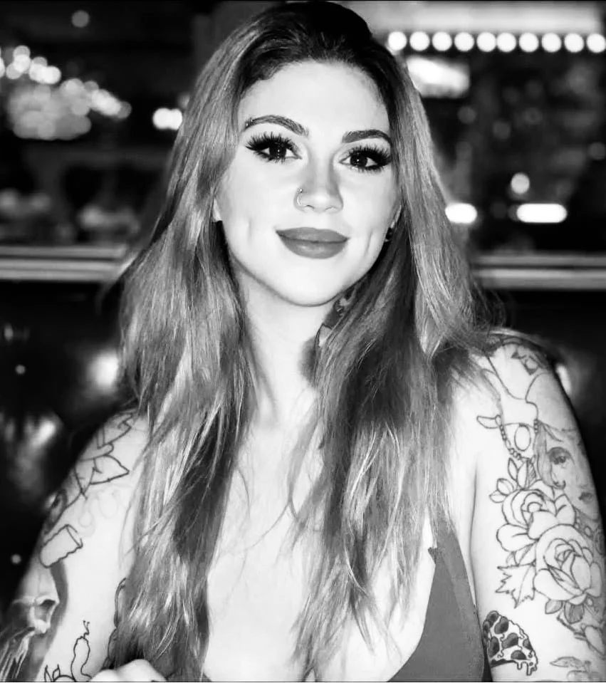 Black and white photo of a young woman with long wavy hair, tattoos on her arms, and a nose ring, smiling in a dimly lit environment with blurry lights in the background.
