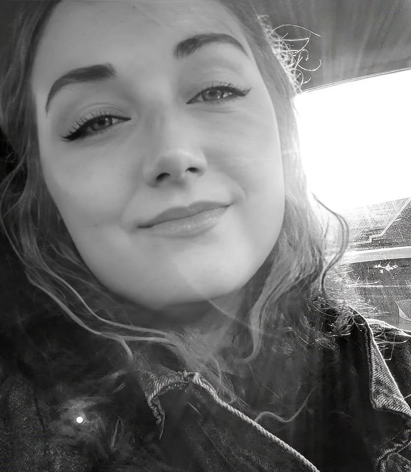 A black-and-white close-up photo of a smiling woman with wavy hair, wearing a denim jacket, inside a car with sunlight coming through the window.