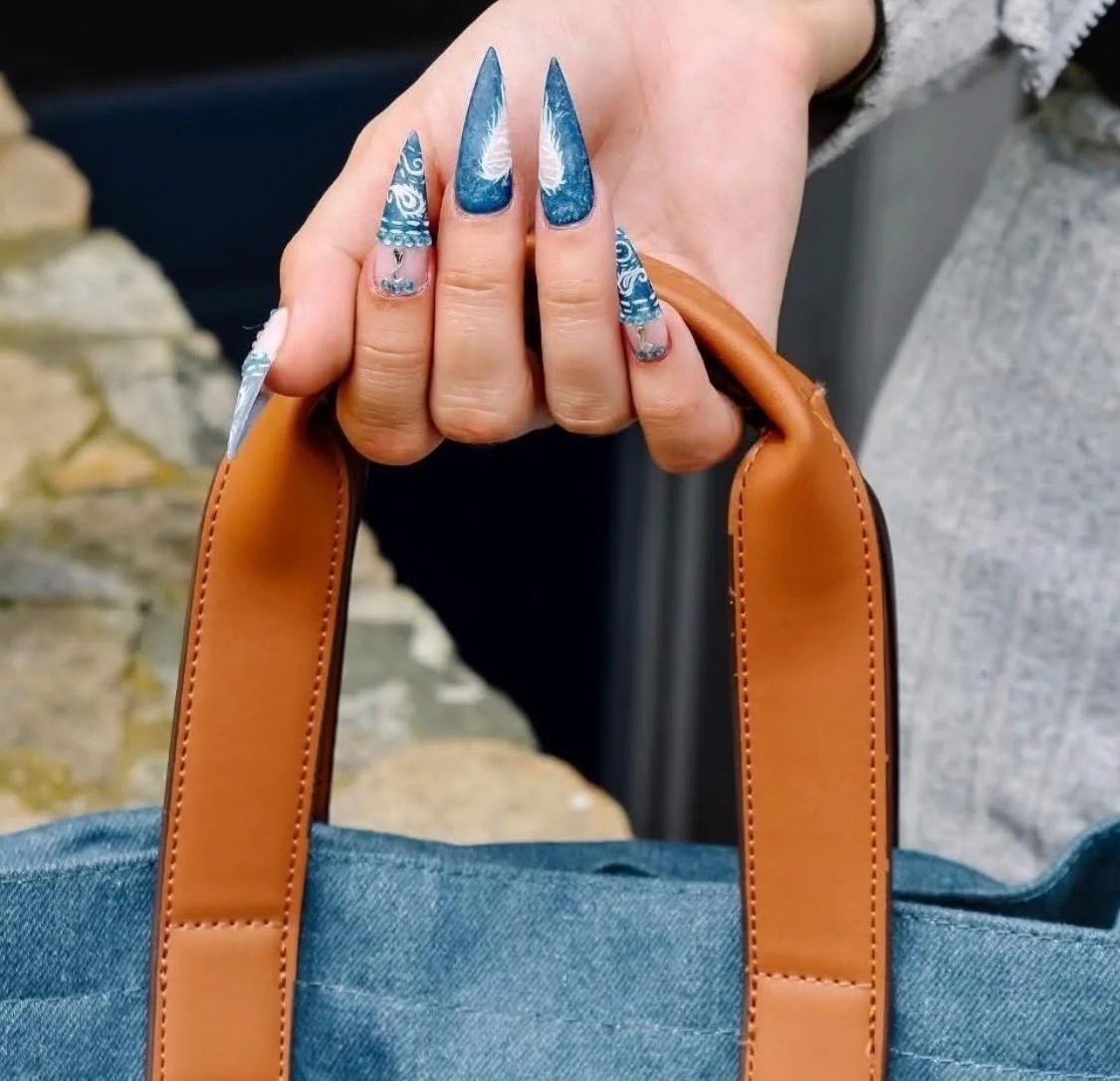 A close-up of a person's hand with long, decorated, blue patterned acrylic nails holding onto tan leather handles of a bag. The person appears to be wearing a gray and black clothing. The background includes a stone surface and part of a gray wall.