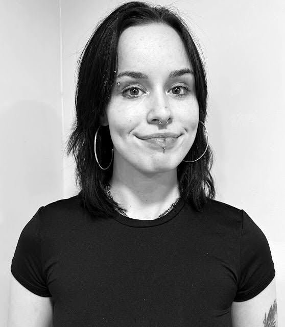 A young woman with dark hair, multiple facial piercings, and large hoop earrings, smiling in black and white.