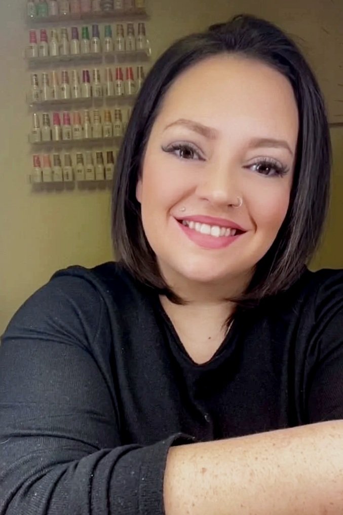 A woman with shoulder-length dark hair smiling in a nail salon, with nail polish bottles displayed on wall shelves behind her.