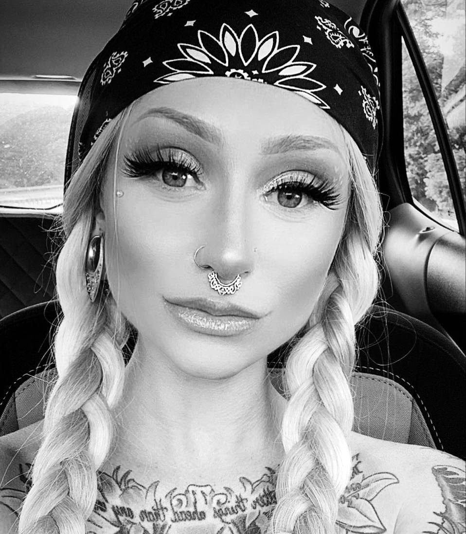 A young woman with long braided hair, wearing makeup, a bandana on her head, and large hoop earrings. She has multiple facial piercings, including a septum ring and nose stud. She is sitting in a car with trees visible through the window in the background.