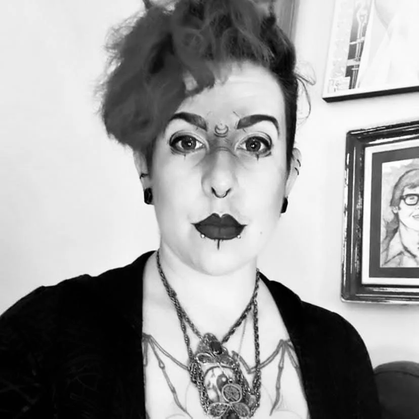 Black-and-white portrait of a person with short, styled hair, wearing dark lipstick, eye makeup, and multiple necklaces, standing in front of a wall with framed artwork.