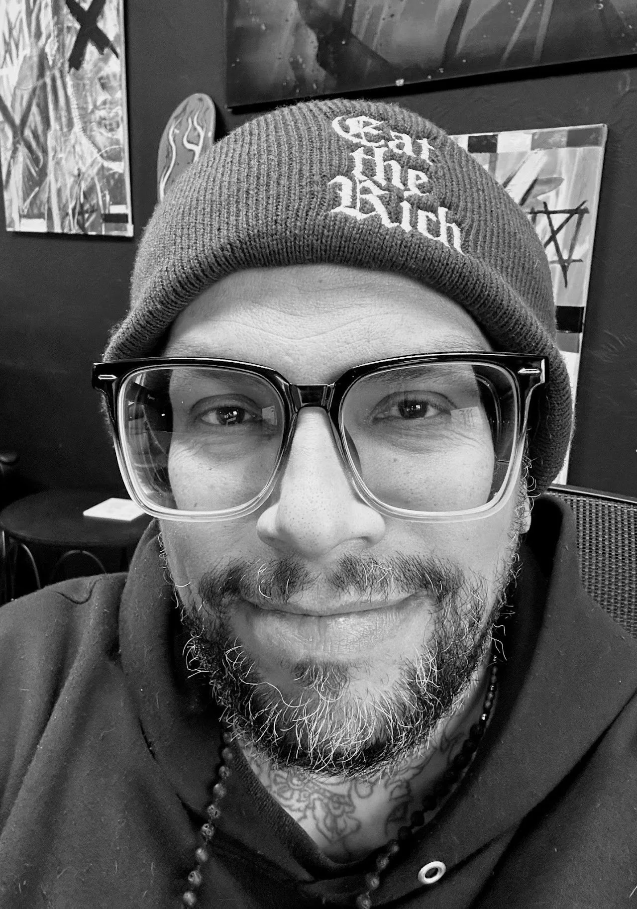 A man with glasses and a beanie taking a selfie in a room with artwork on the wall behind him.